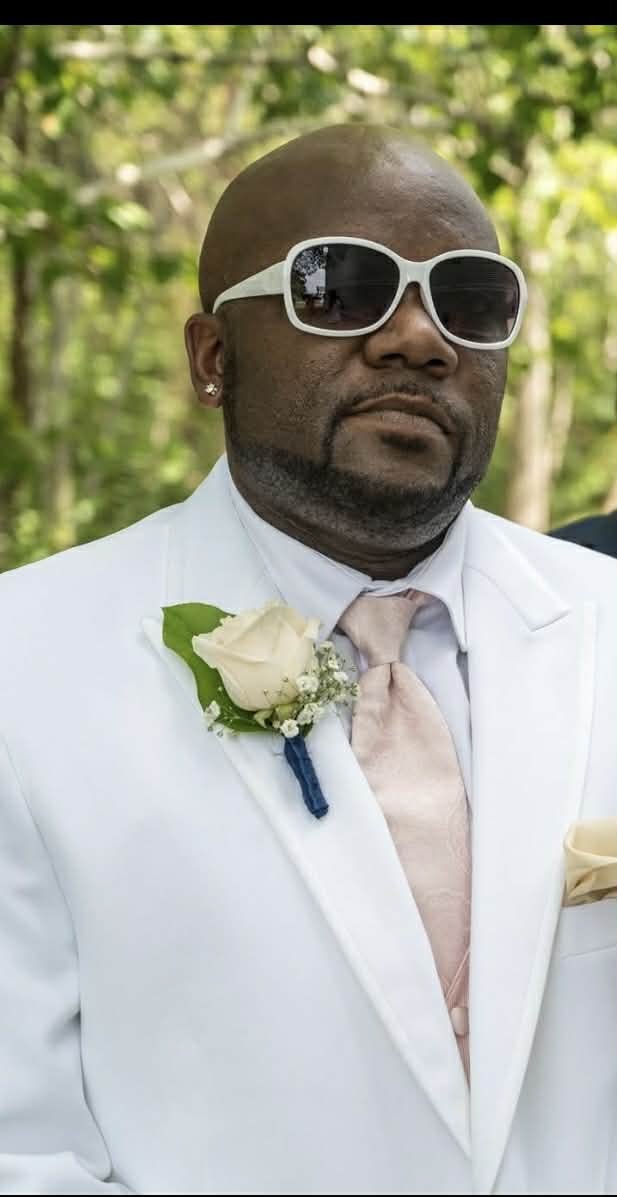 A man wearing a white suit, pink tie, white sunglasses, and a white boutonniere, standing outdoors with green foliage in the background.