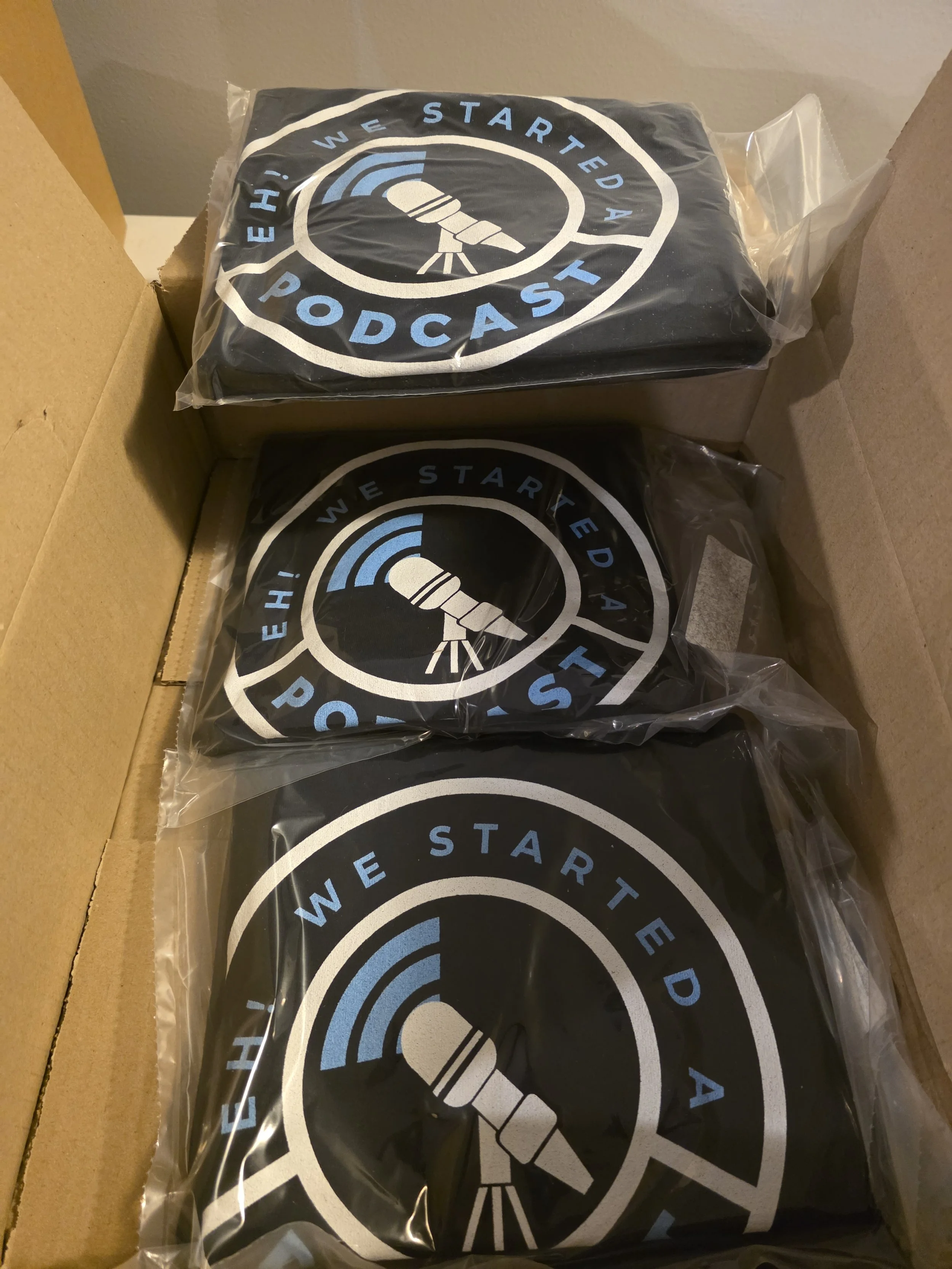Three black t-shirts with the logo of the "EH! We Started a Podcast" show, featuring a stylized telescope, inside a cardboard box, wrapped in clear plastic.