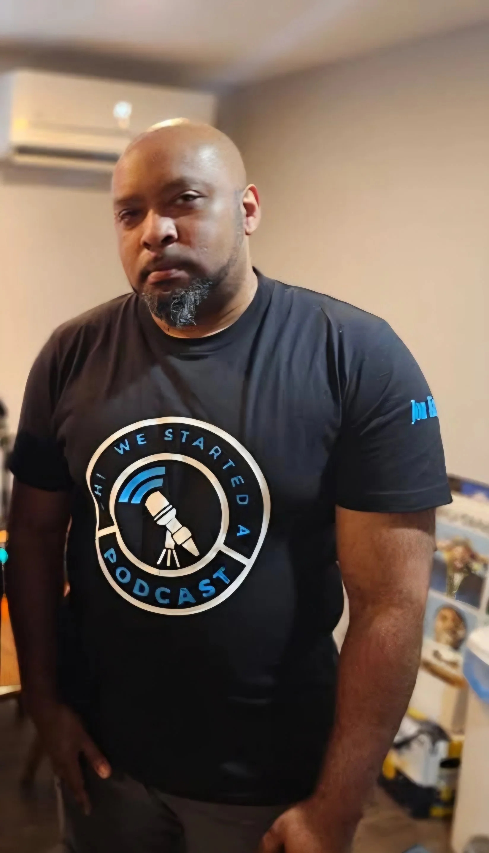 A man with a bald head and beard wearing a black T-shirt with a podcast logo that says 'We Started A Podcast' and a microphone graphic, standing indoors.