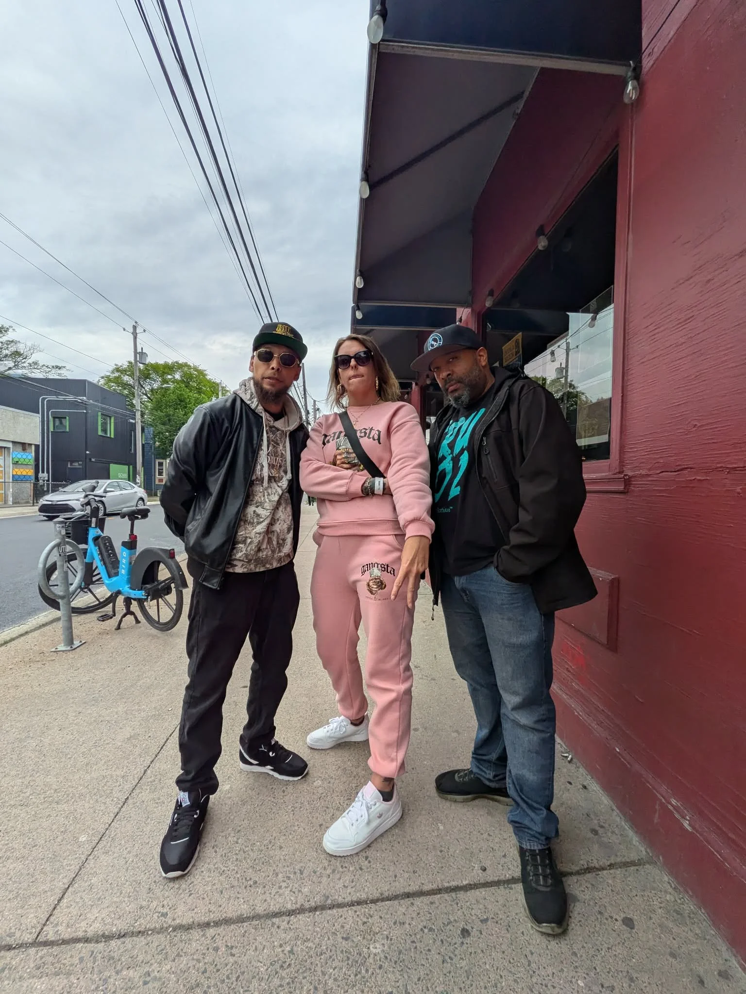 Three people standing on the sidewalk outside a building, with a red wall to their right. The person on the left wears sunglasses, a black hat, a black jacket, and black pants. The person in the middle is a woman with shoulder-length hair, sunglasses