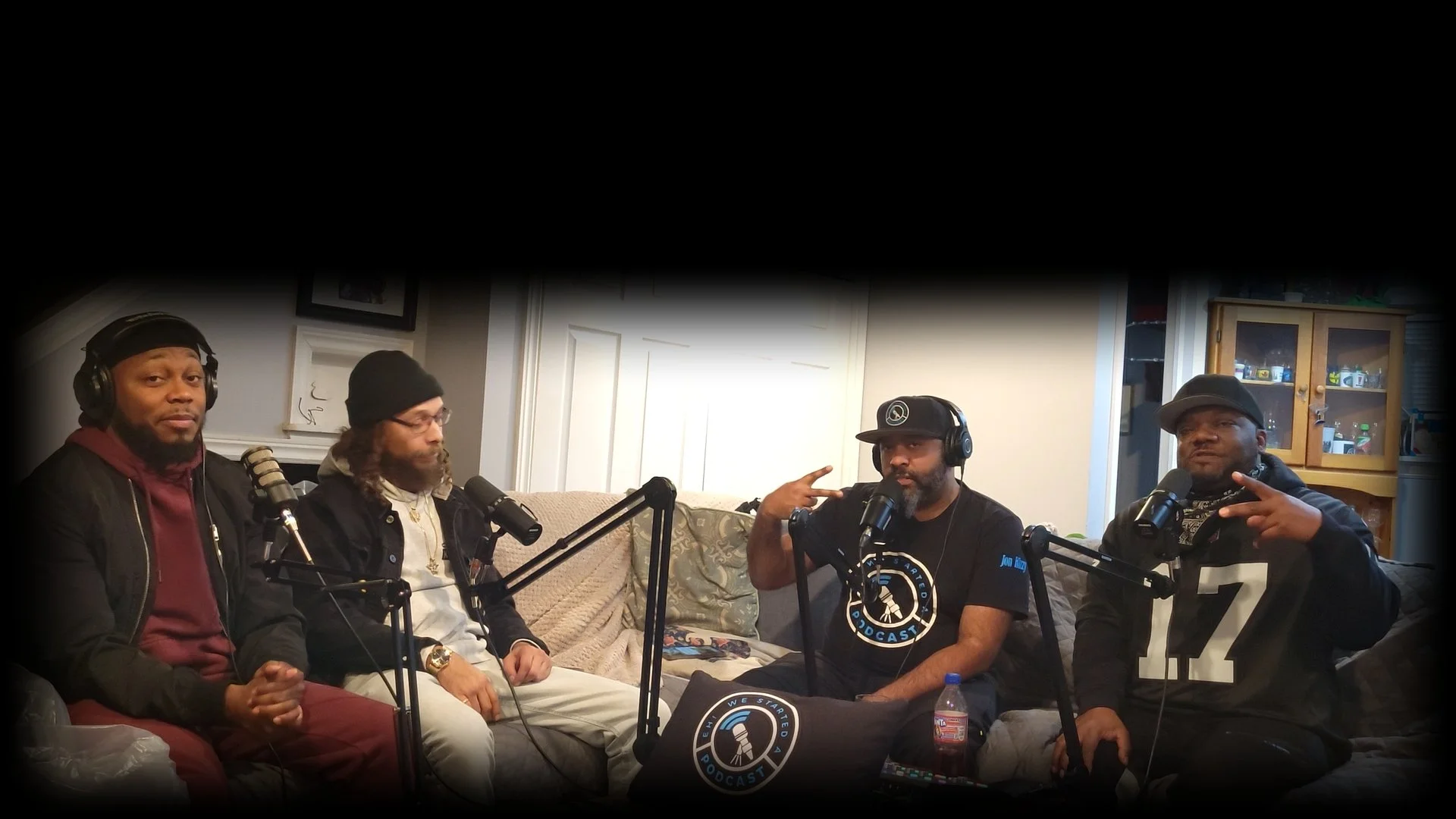 DJ Laton, Ray Rey, Jon Kizzy and King Tygga sitting on a beige couch and chairs with microphones in front of them, recording a podcast in a cozy room with cabinets and pictures on the wall.