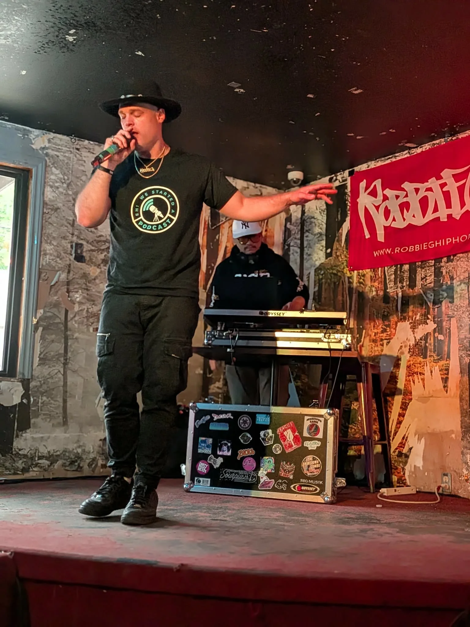 A man wearing a black hat, black t-shirt with a graphic and the words 'We Started a Podcast', and black cargo pants singing into a microphone on stage. Behind him, a DJ or music producer wearing a baseball cap and hoodie managing equipment. The backg