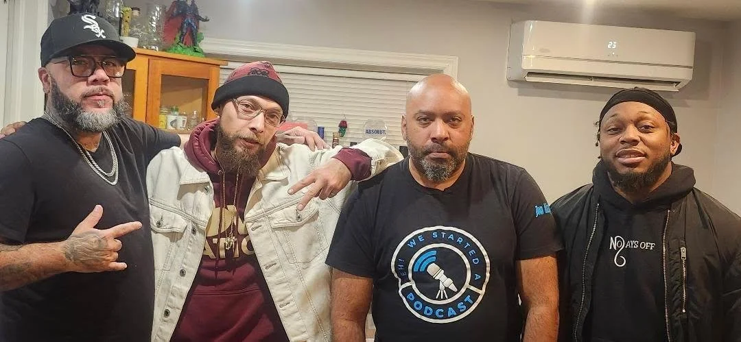 Group of five men standing indoors, posing for a photo. The man on the left wears glasses, a black baseball cap, and a black t-shirt. The second man wears glasses, a black beanie, and a cream-colored jacket. The middle man is bald, with a beard, wear