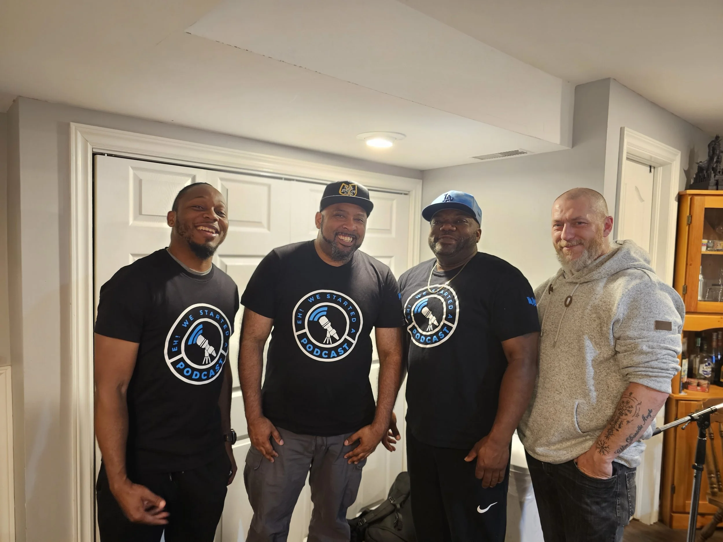 DJ Laton, Jon Kizzy, King Tygga and Shane standing side by side indoors, all smiling, wearing black shirts with a podcast logo, and one man in a grey hoodie with tattoos on his arms.