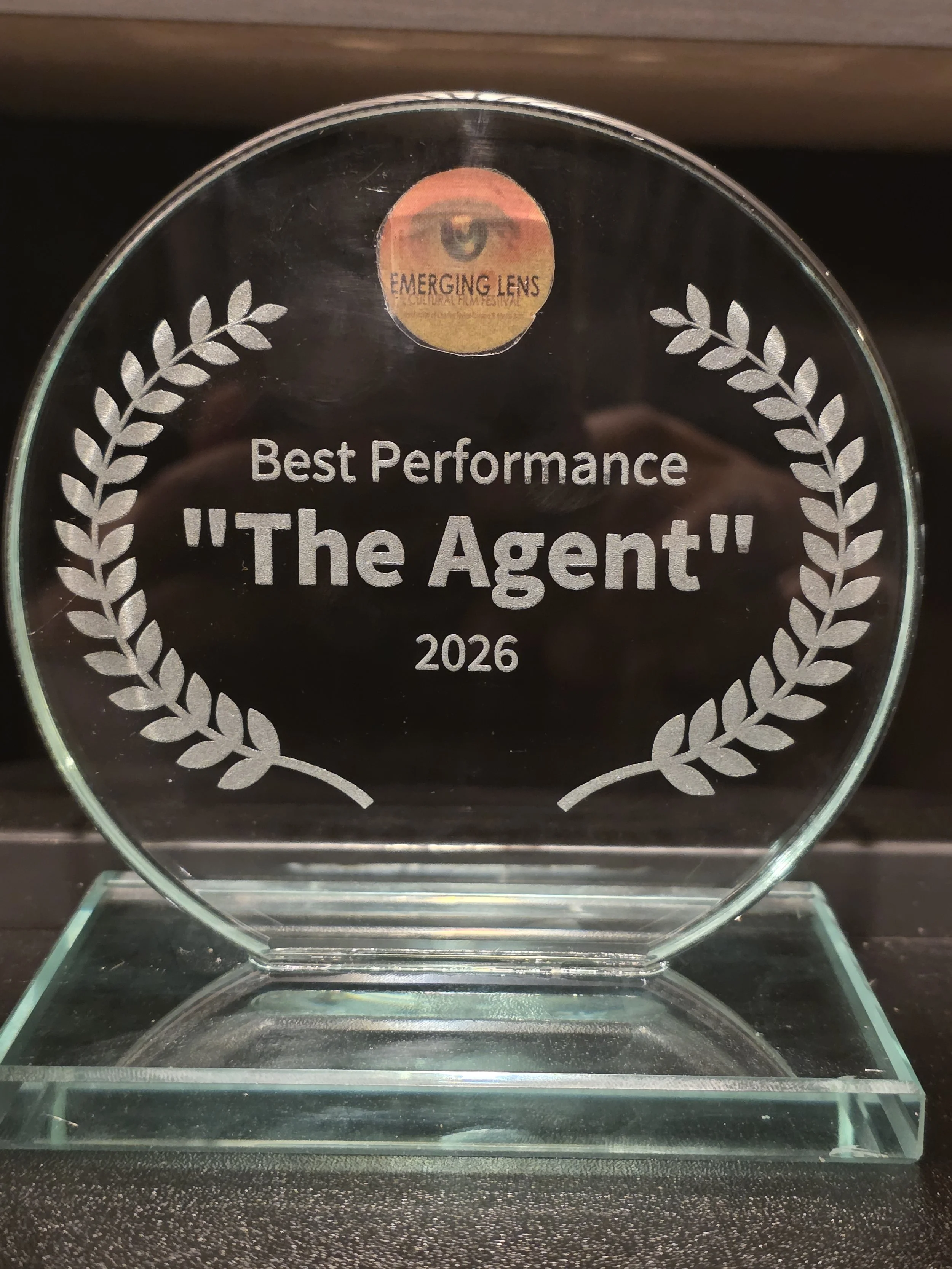 A glass award with laurel leaves engraved on it, inscribed with 'Best Performance "The Agent" 2026', from the Emerging Lens Cultural Film Festival.