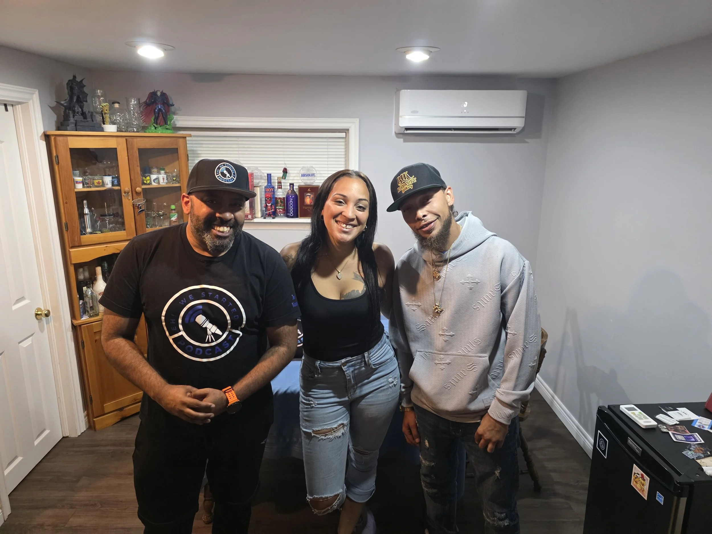 Jon Kizzy, Realeyes and Ray Rey smiling and posing indoors, with a wooden cabinet and decorations including figurines and bottles behind them.
