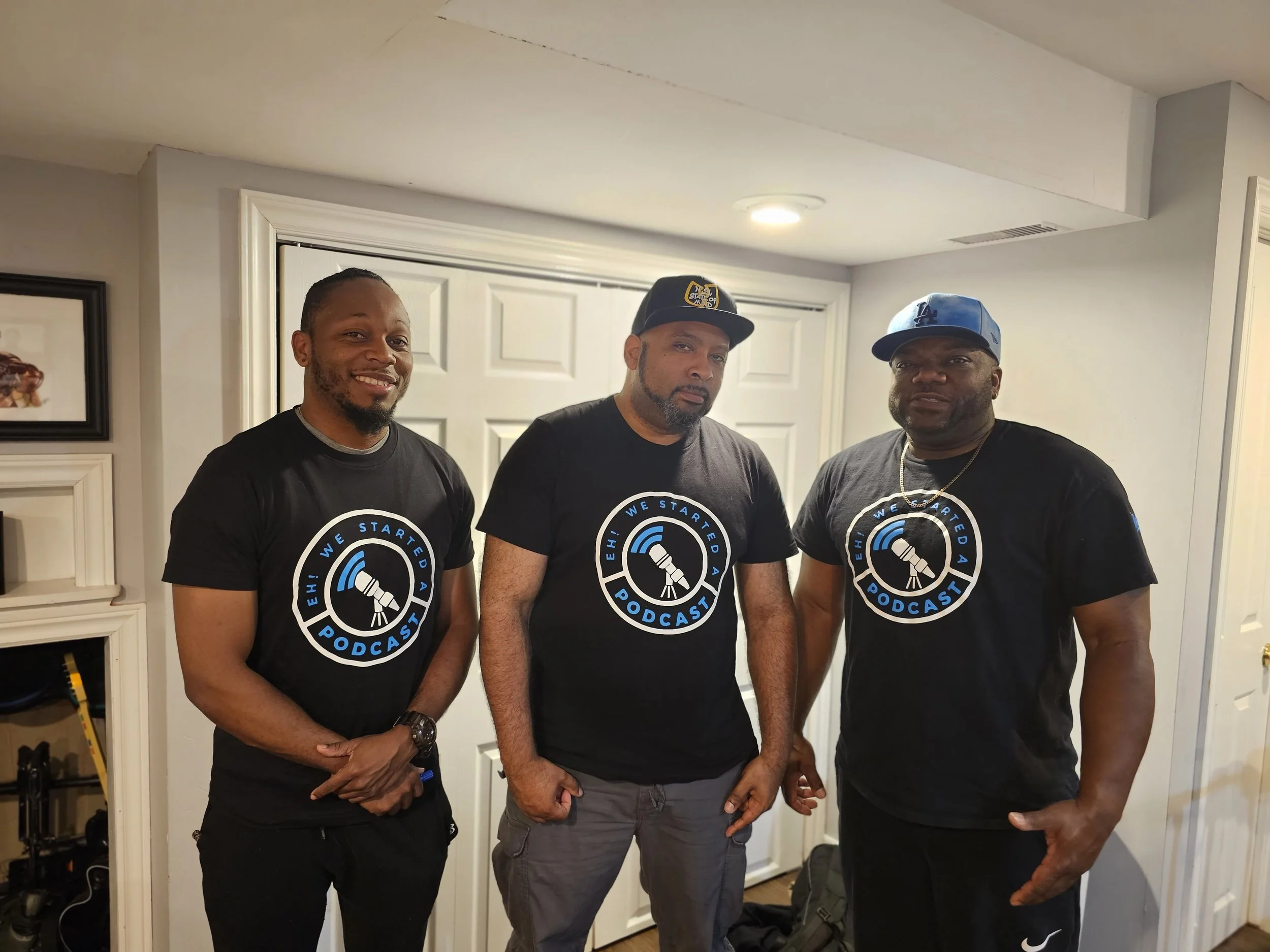 Three men standing together indoors, all wearing black t-shirts with a podcast logo that says 'We Started A Podcast' with a graphic of a microphone and Wi-Fi symbol. The man on the left is smiling with his hands clasped, the middle man has a serious expression with a black cap, and the man on the right is looking at the camera wearing a blue cap.