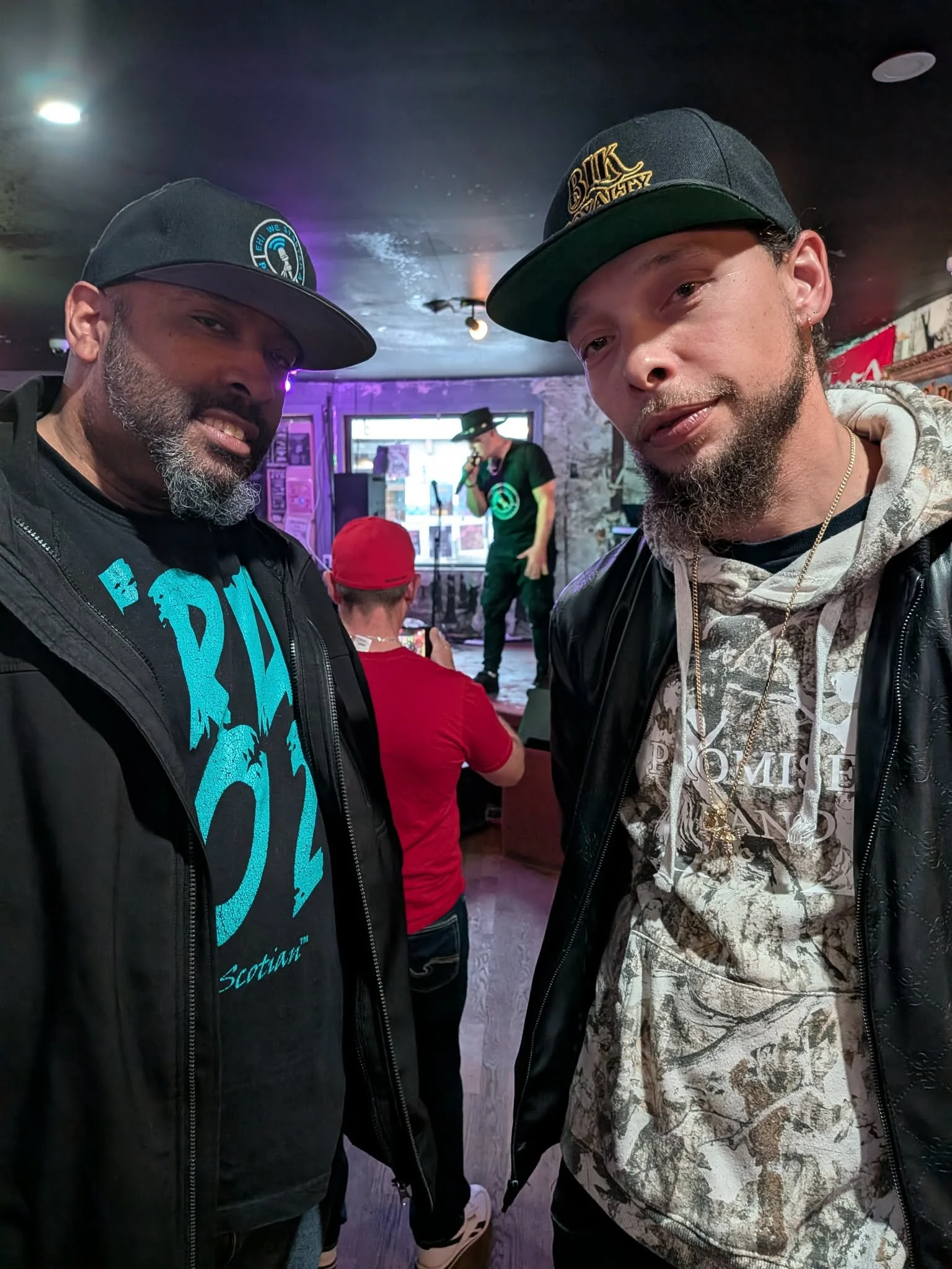 Jon Kizzy and Ray Rey taking a selfie in a dimly lit bar or music venue. Both wearing black caps and jackets. A performer with a microphone is visible in the background, standing on a small stage with a stone wall behind him.