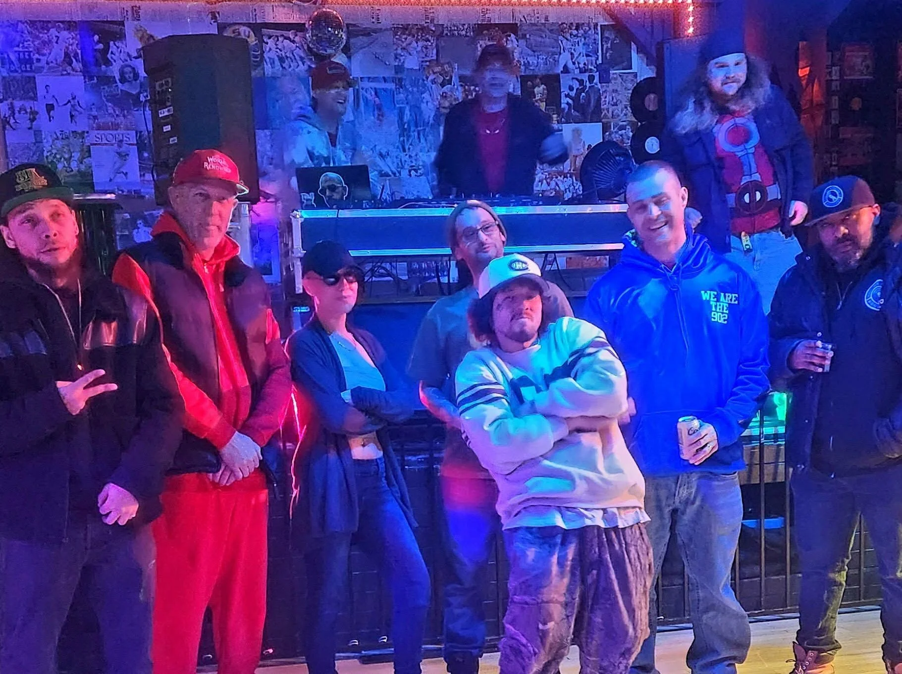 Group of people at a bar or club, some with drinks, standing in front of a colorful, neon-lit background. A woman is wearing a hat with a Deadpool logo, and others are dressed casually; some are smiling and posing for the photo.