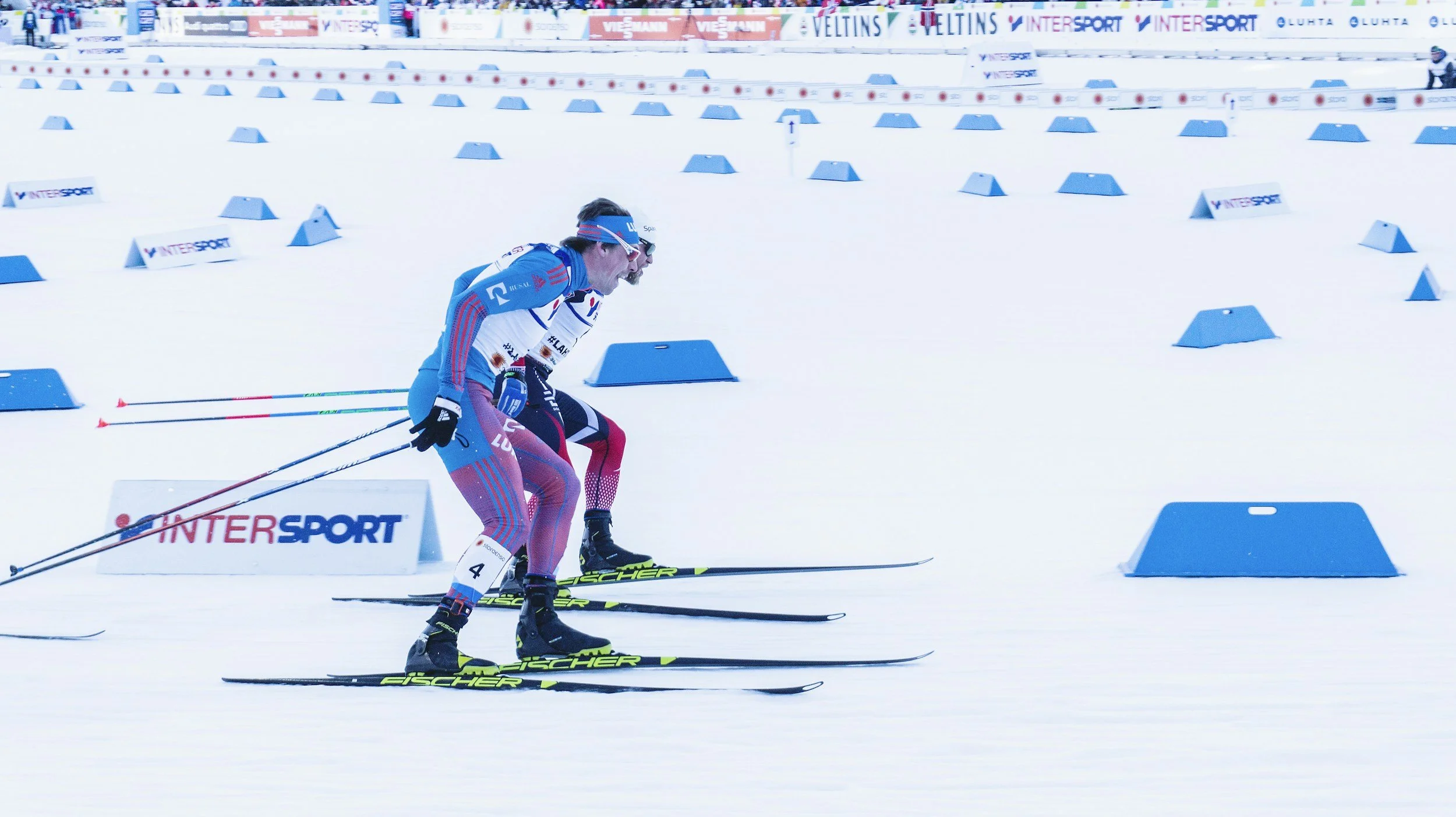 A cross-country skier wearing a blue, white, and red outfit and a headband, pushing off on snow covered course marked with blue barriers and 'INTERSPORT' banners, during a race.