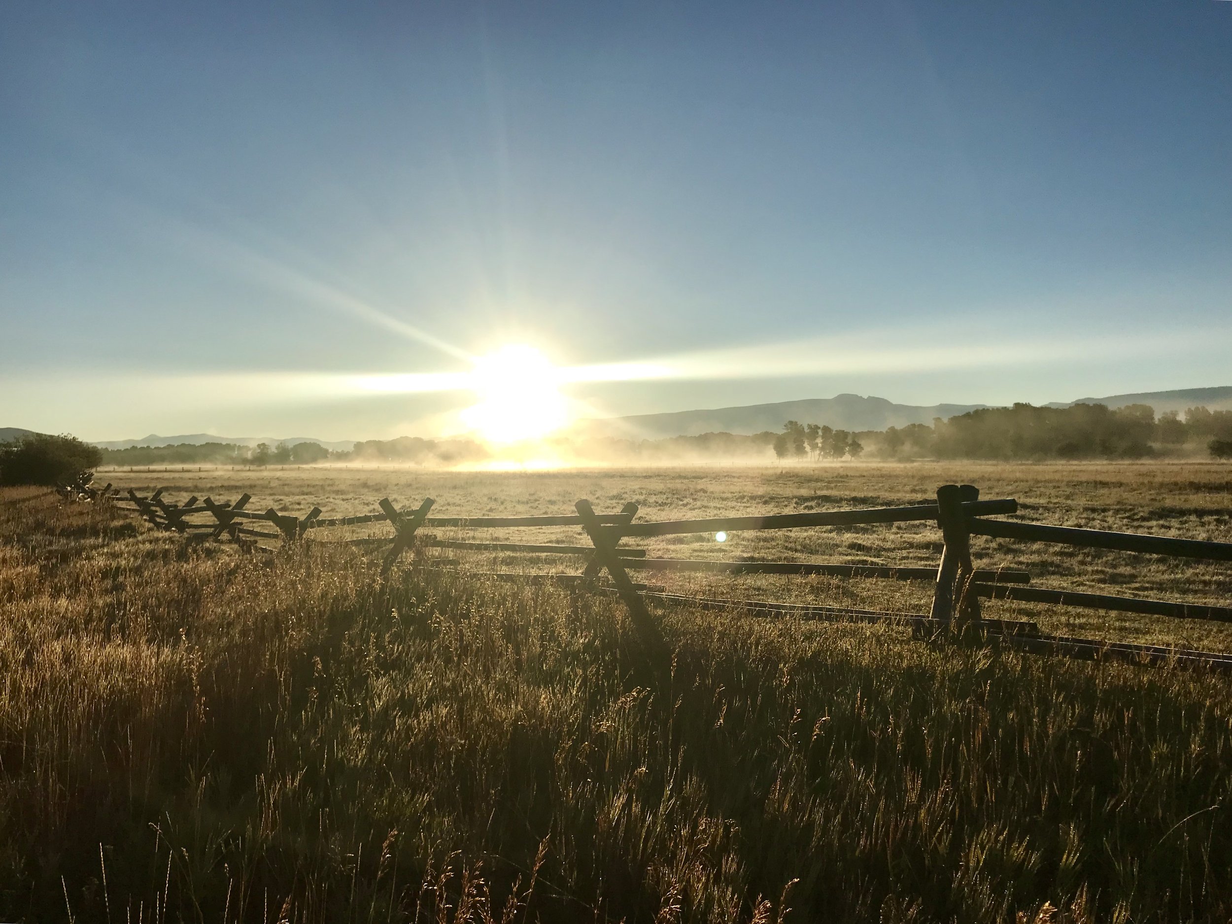 Sunrise over a farm field with a wooden fence in the foreground and distant hills on the horizon.
