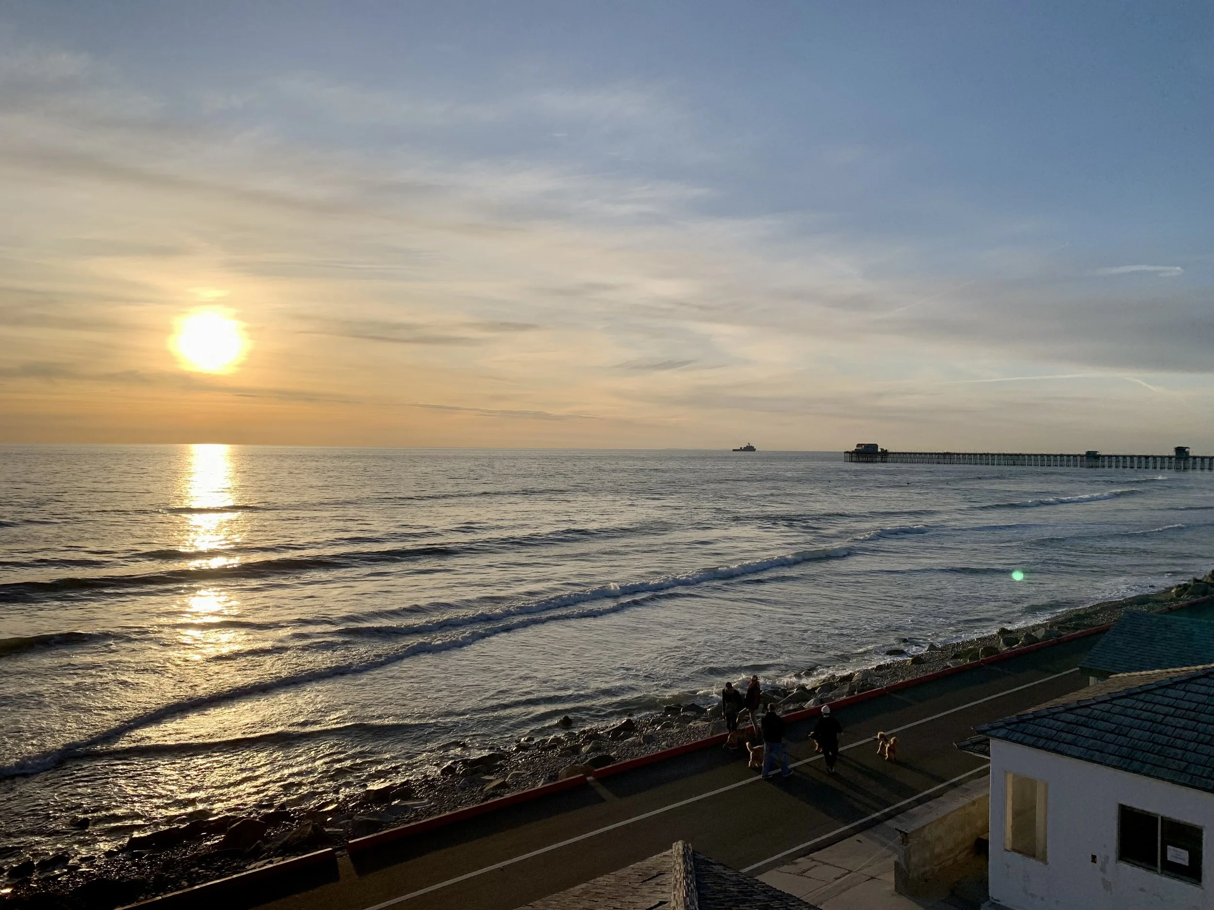 Sunset over the ocean with a pier extending into the water, a few buildings on the shoreline, and people walking along a rocky beach.