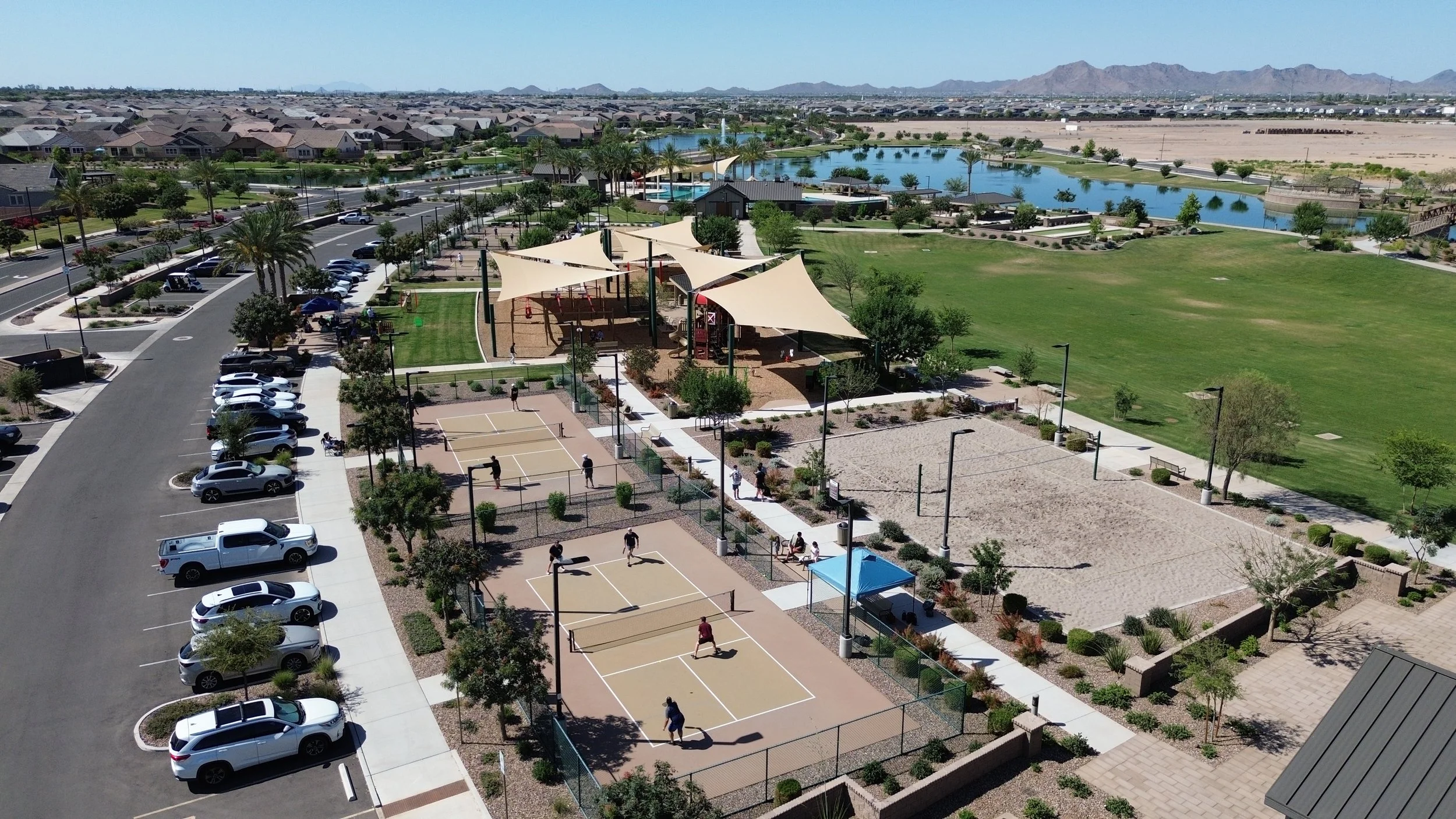Aerial view of a community park featuring multiple tennis courts with players, a playground with canopy shades, a large grassy area, a swimming pool with a covered pavilion, a pond, and adjacent residential houses in the background. Mountains are visible in the distance.