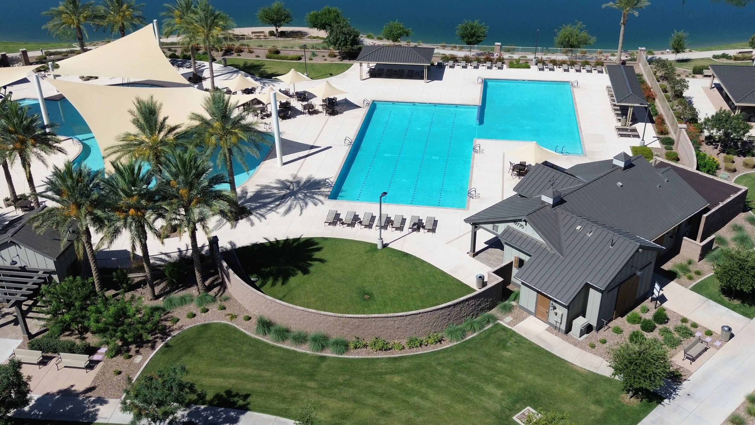 Aerial view of a swimming pool area with lounge chairs, shade structures, palm trees, and a small clubhouse, overlooking a body of water.