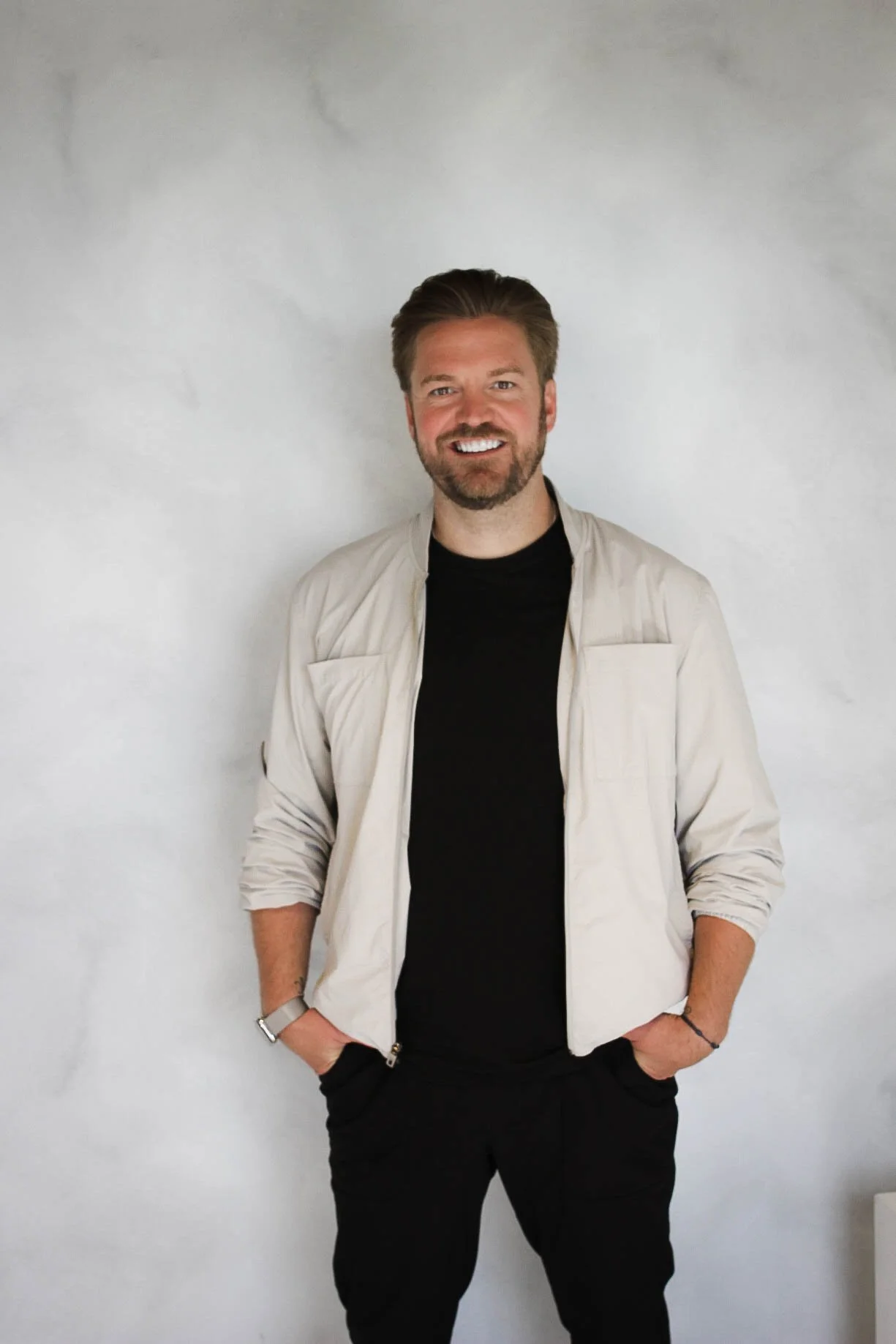 A smiling man with a beard and styled hair standing against a light-colored wall, wearing a black t-shirt, a beige jacket with rolled-up sleeves, black pants, and a watch on his left wrist.