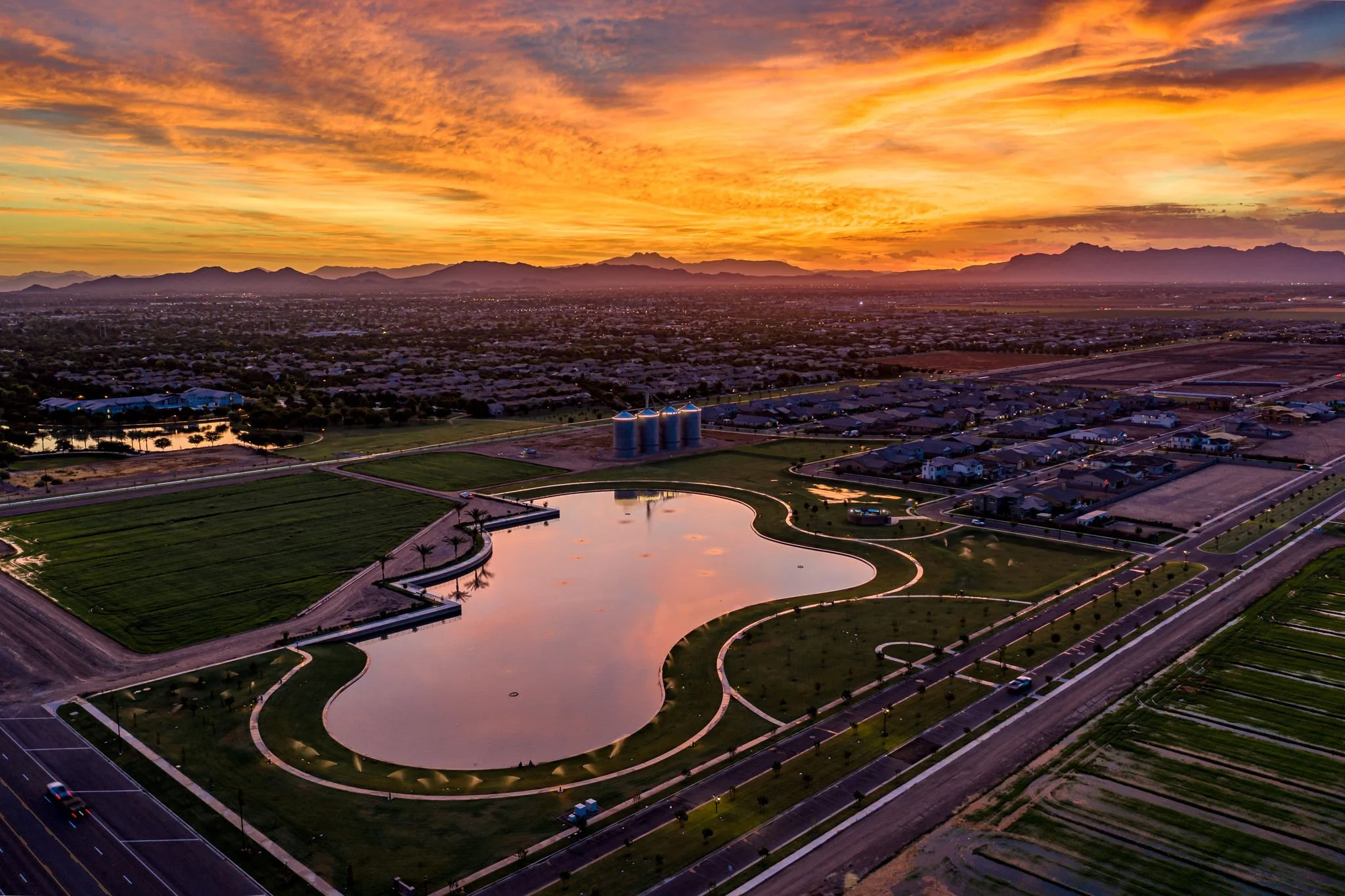 Aerial view of a city at sunset with mountains in the background, a large, reflective pond with a curved shape in the foreground, and a farm with silos and fields nearby.