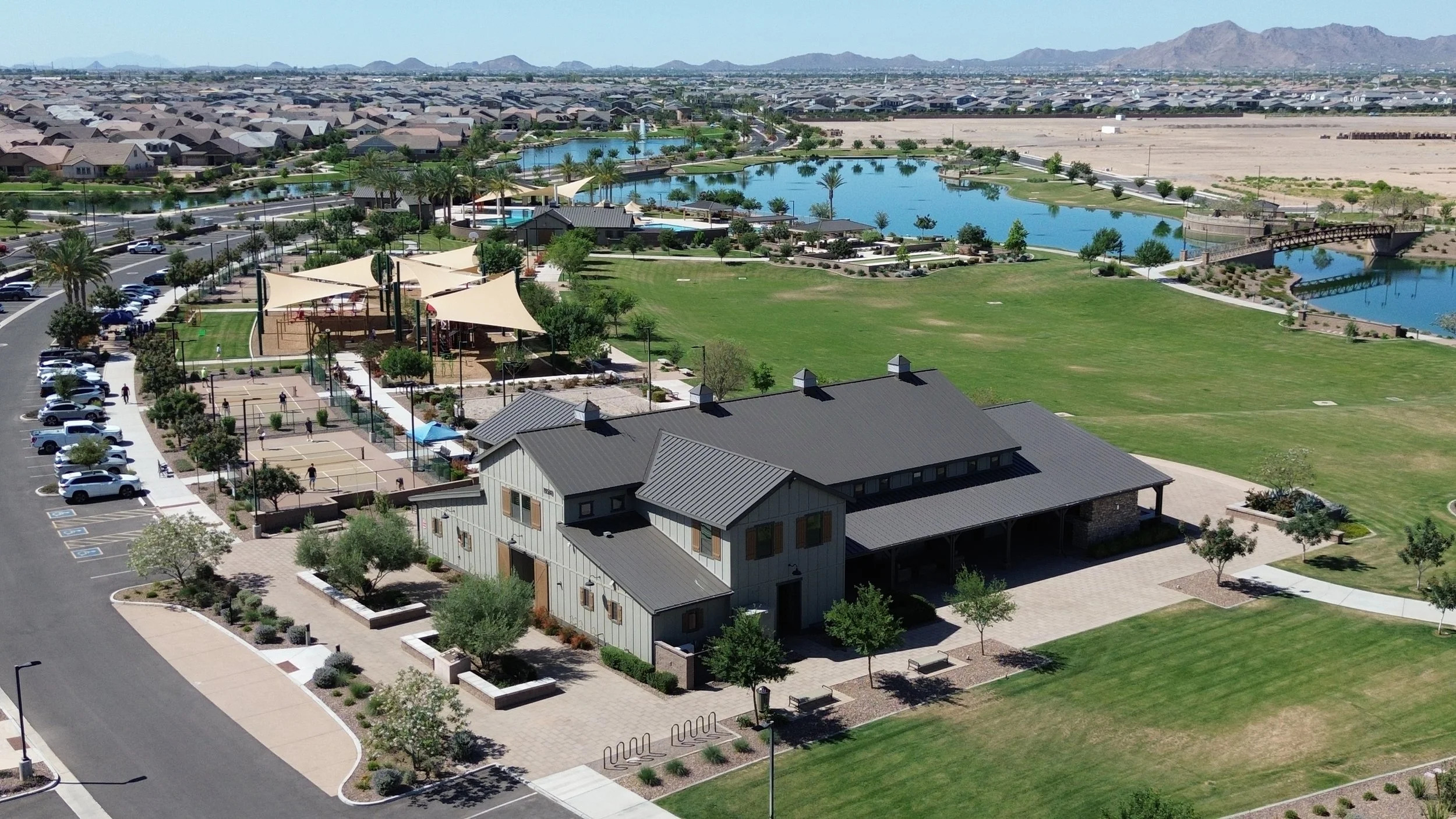 Aerial view of a suburban park with lakes, walking paths, playground, picnic areas, tennis courts, parking lot, residential houses, and mountain range in the background.