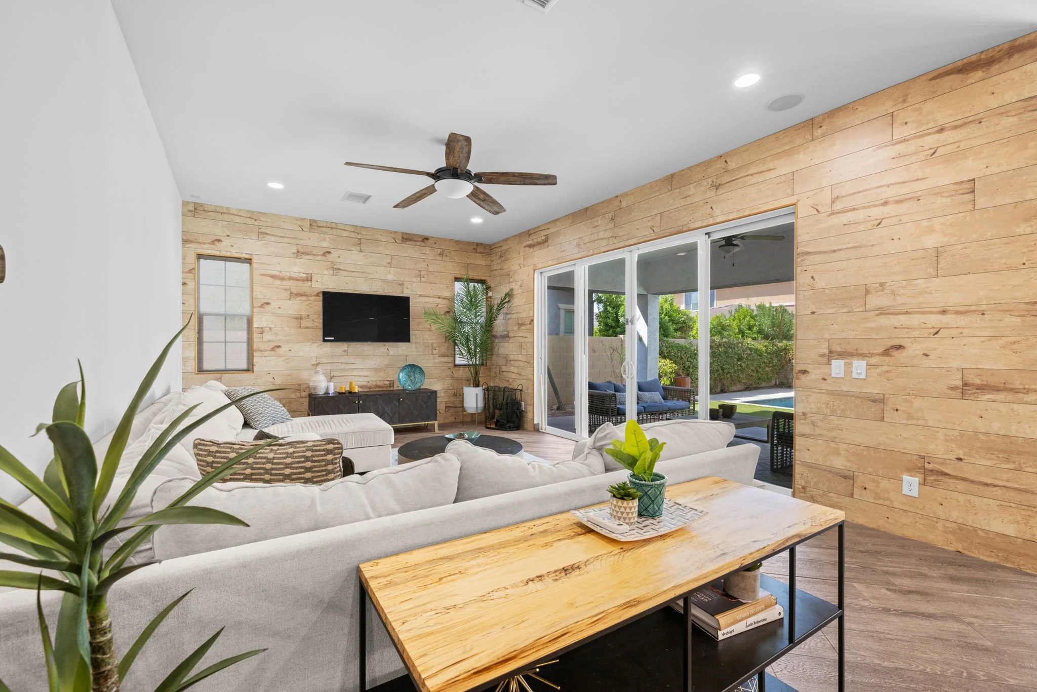 A modern living room with a light-colored sectional sofa, a wooden coffee table, and a black media console with a flat-screen TV. The room features a wooden accent wall, large glass sliding doors leading outside, potted plants, and a ceiling fan with wooden blades.