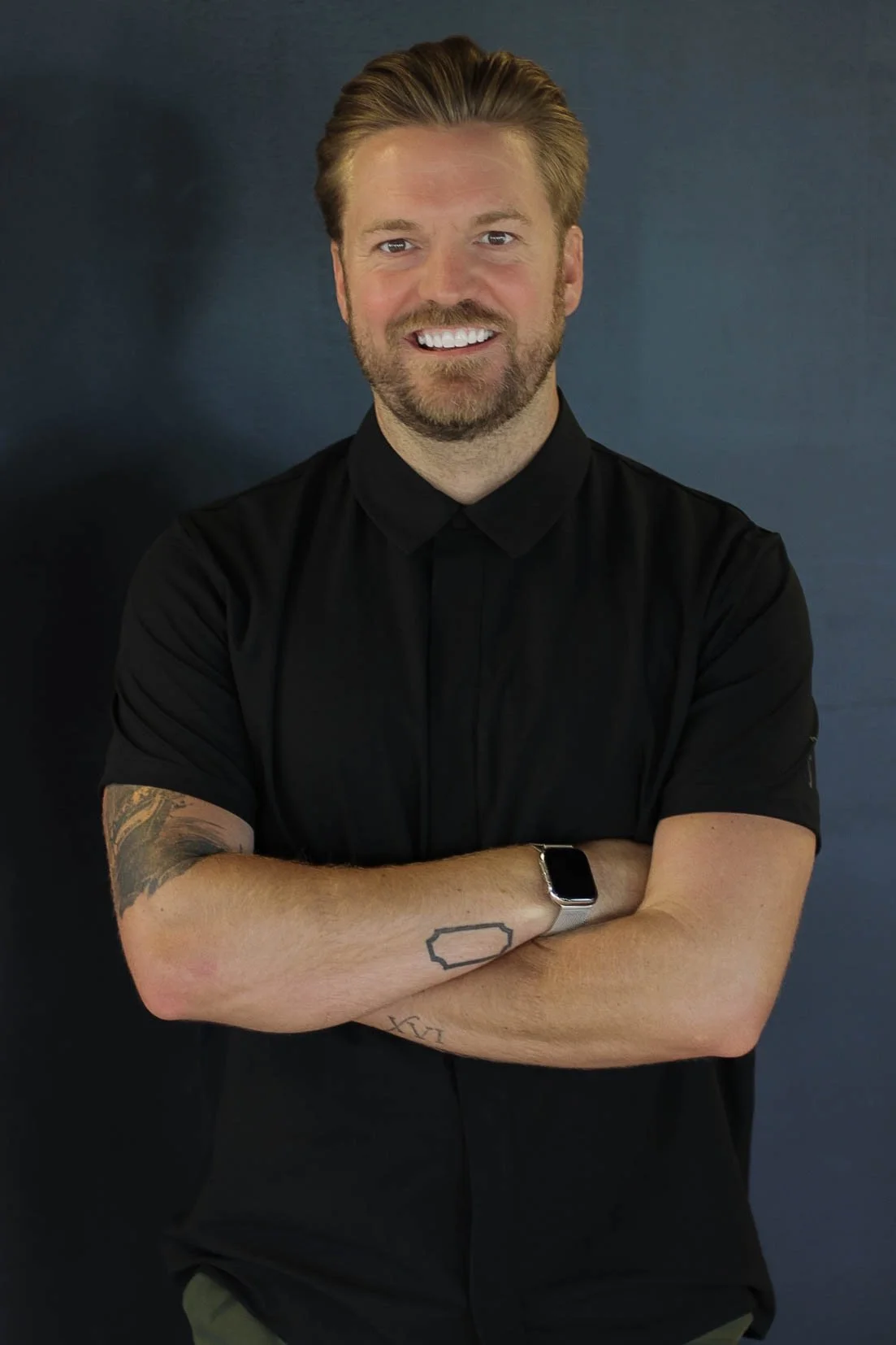 A man with light brown hair and a beard, smiling, wearing a black shirt and a smartwatch, standing against a dark background.