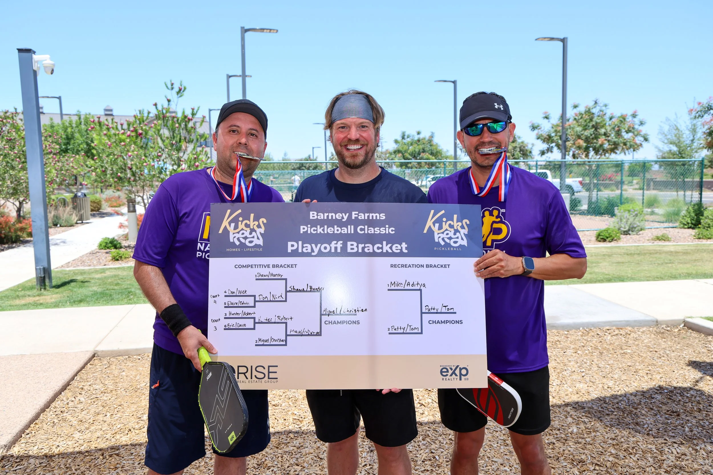 Three men standing outdoors holding a bracket for a pickleball tournament, smiling and biting medals, with a playground and trees in the background.