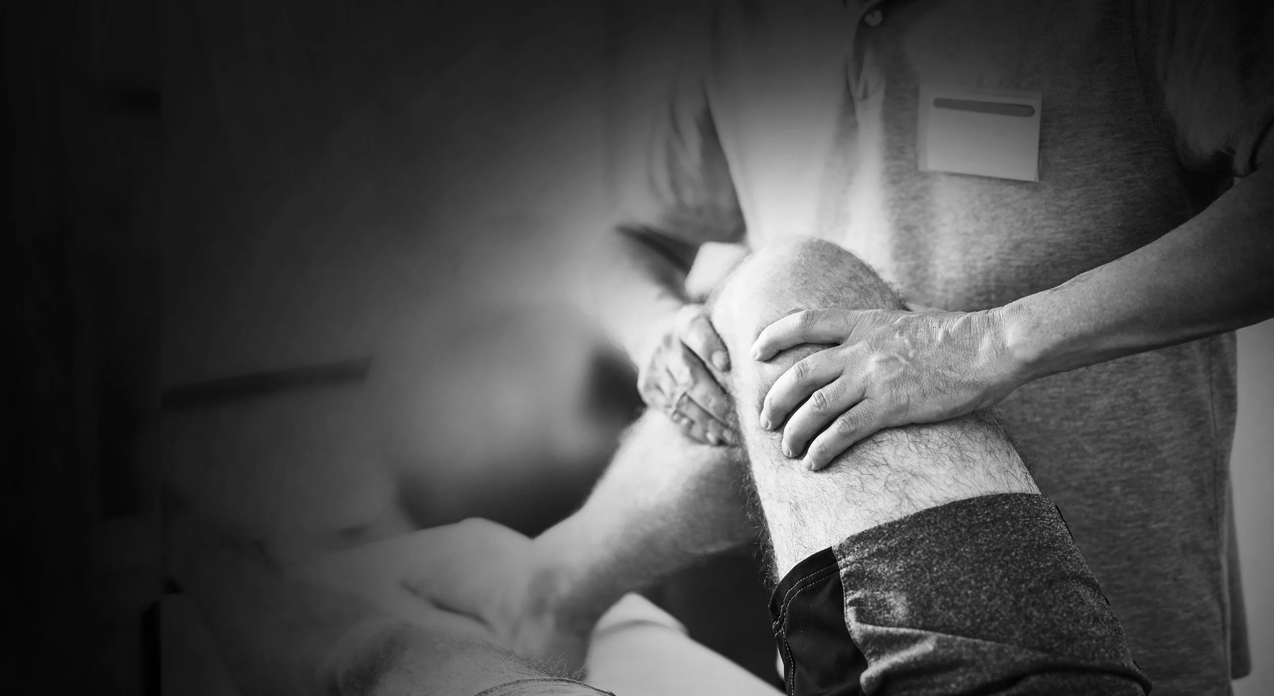 Patient liegt auf einer Behandlungsliege, während ein Therapeut das angewinkelte Bein mit den Händen behandelt. Das Kampagnenmotiv „Gesund werden braucht Nähe“ steht für die Unterstützung der Betriebskrankenkassen bei Rehabilitation und Genesung.