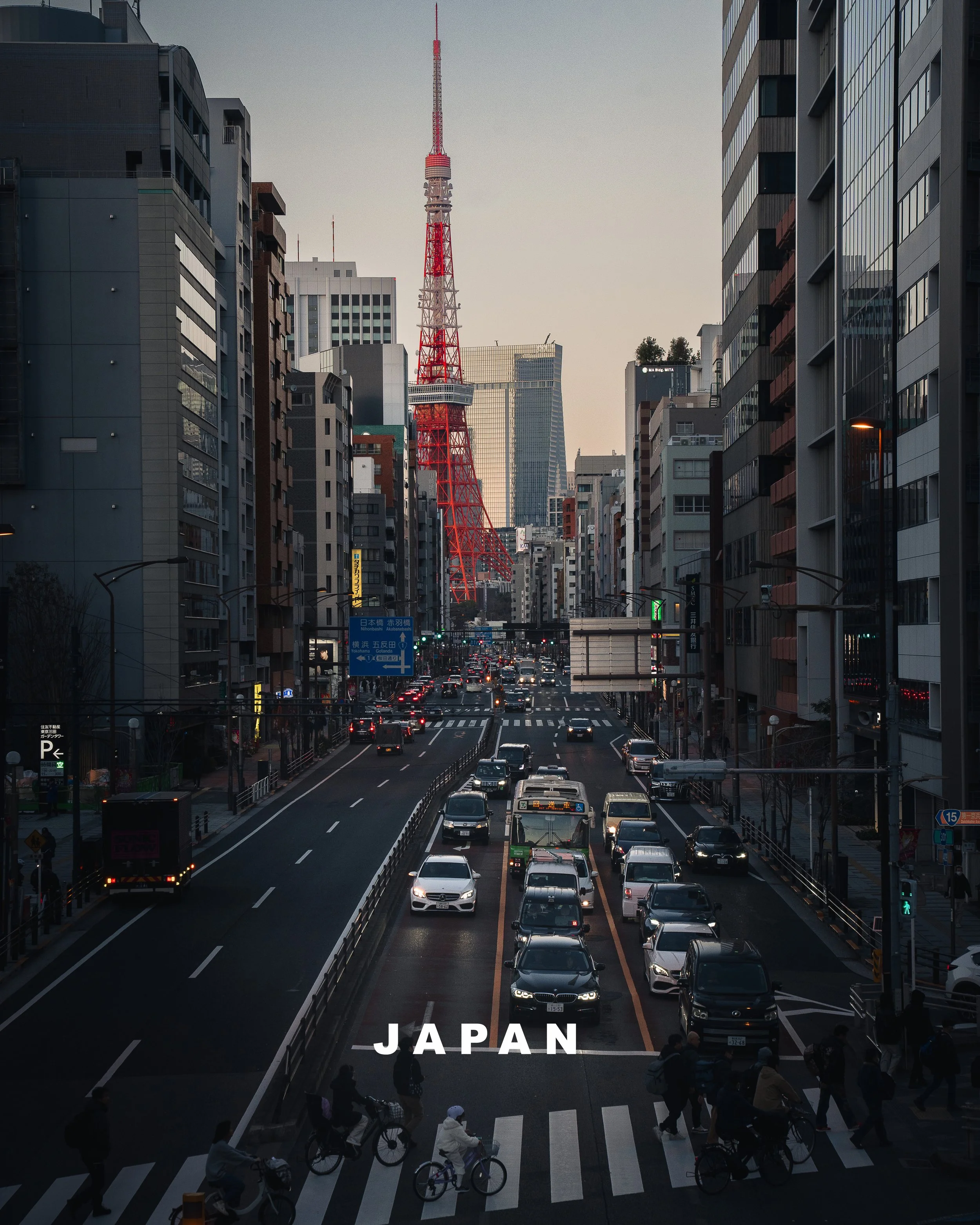 Stadtbild mit Tokyo Tower im Hintergrund, zu sehen sind Mehrfamilienhäuser, Autos auf der Straße und Fußgänger auf dem Zebrastreifen in Japan.