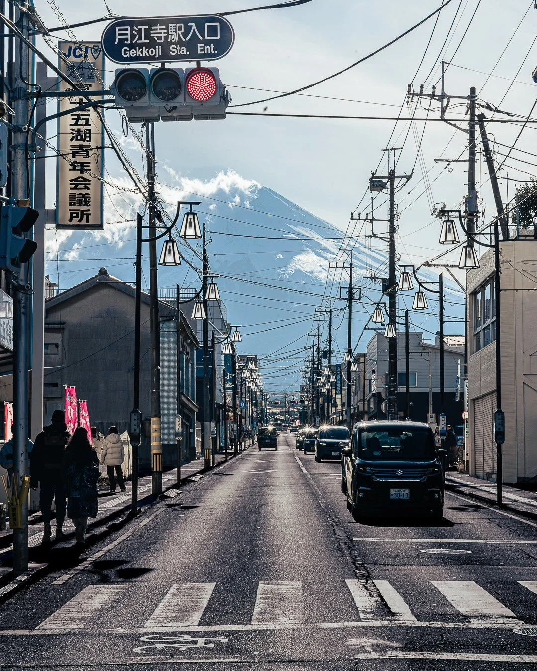 Stadtstraße mit Fußgängern, Autos und Stromleitungen, mit Berg im Hintergrund, Mount Fuji sichtbar, japanische Schilder, Straßenlaternen, klare Wetterbedingungen.