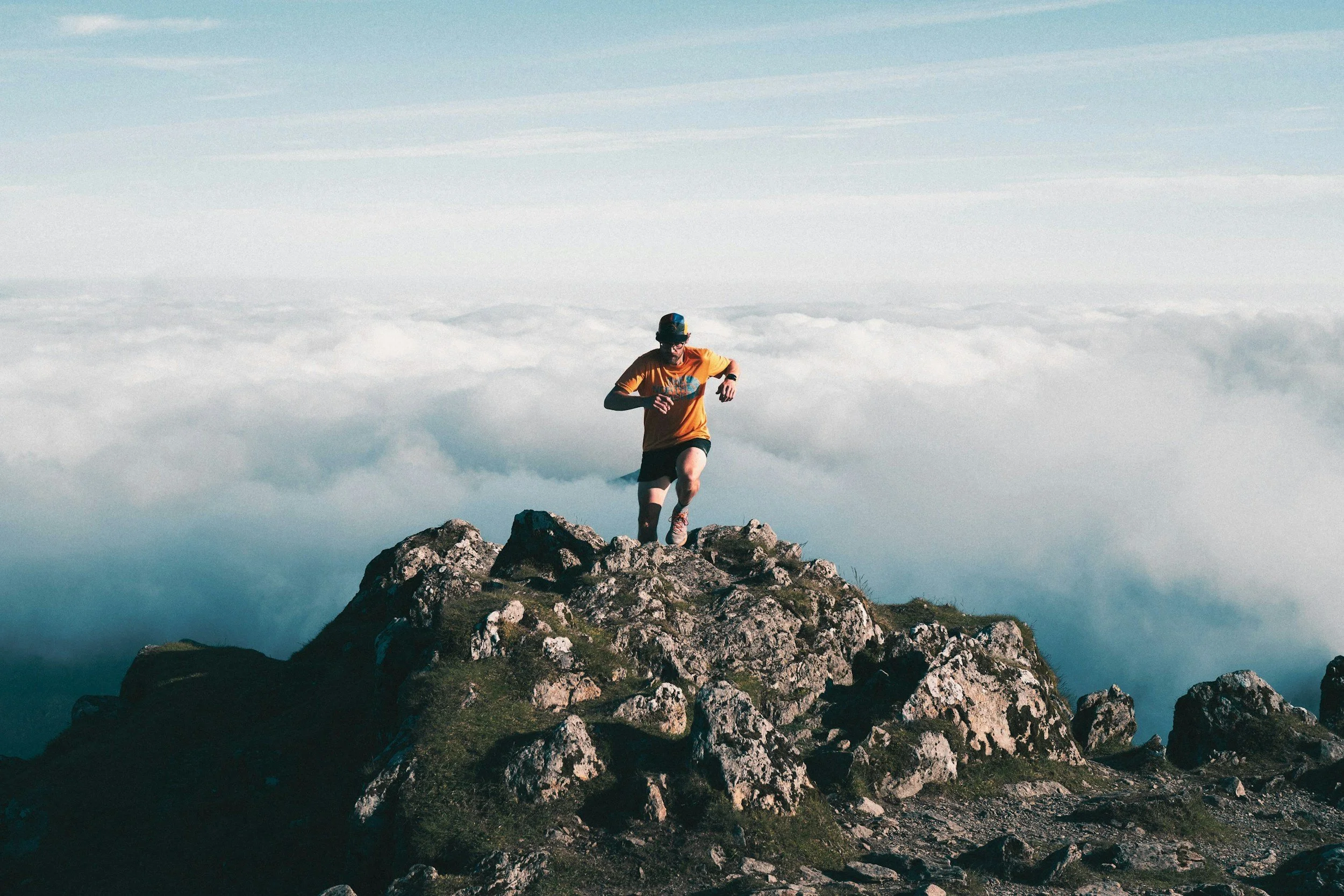 A man running on rocky mountain summit with clouds below and blue sky above.