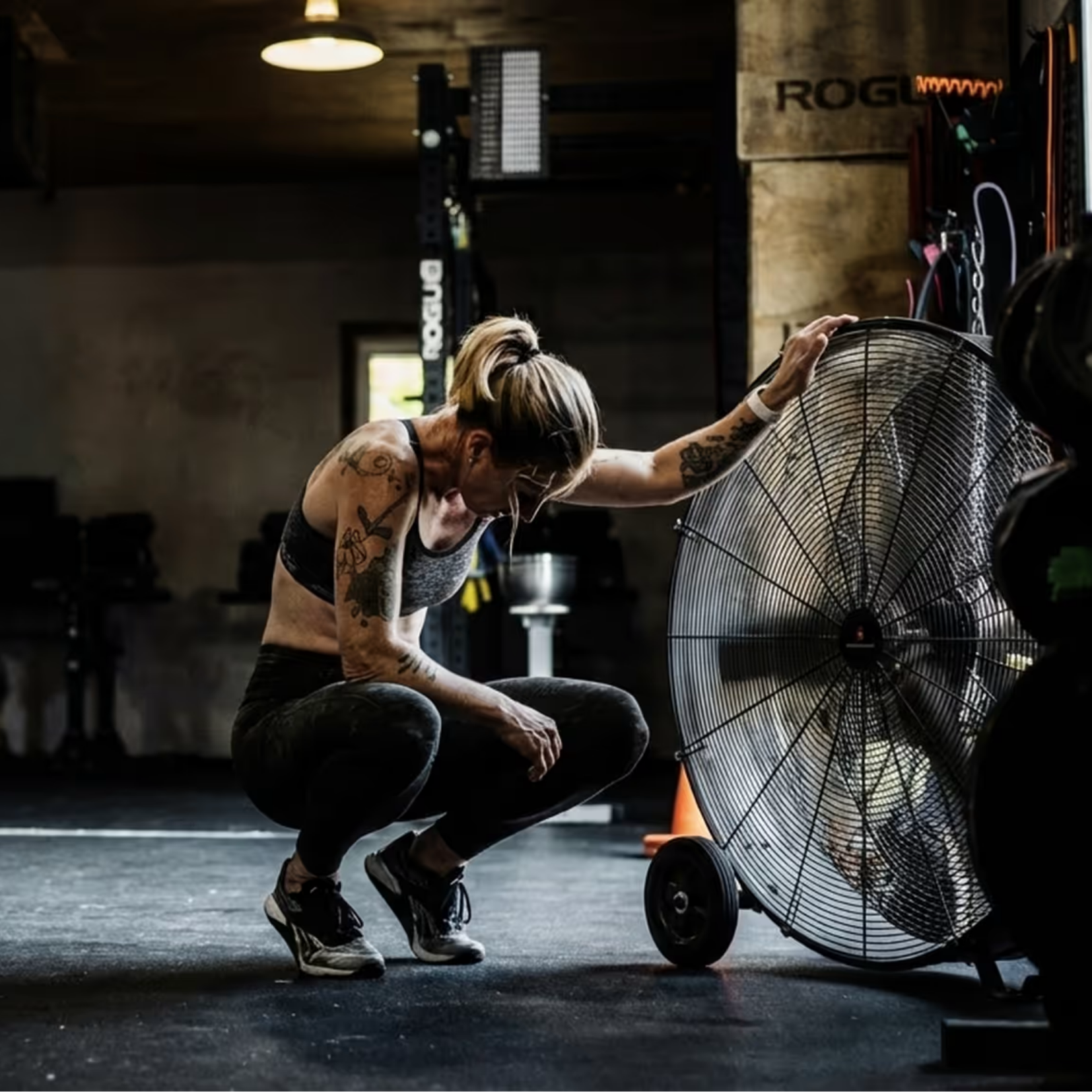 A woman with blonde hair, tattoos, and athletic clothing crouches in a gym, checking her phone near a large industrial fan, with fitness equipment and a ROGUE gym sign in the background.