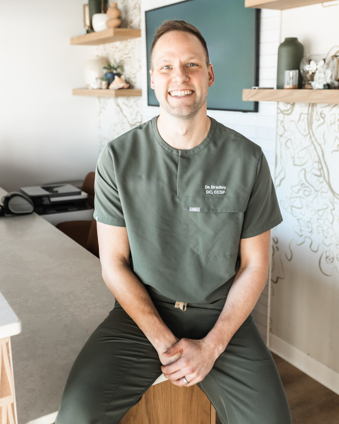 Dr. Bradley Brunner in green medical scrubs sitting on a clinic counter, smiling at the camera in a modern office interior.