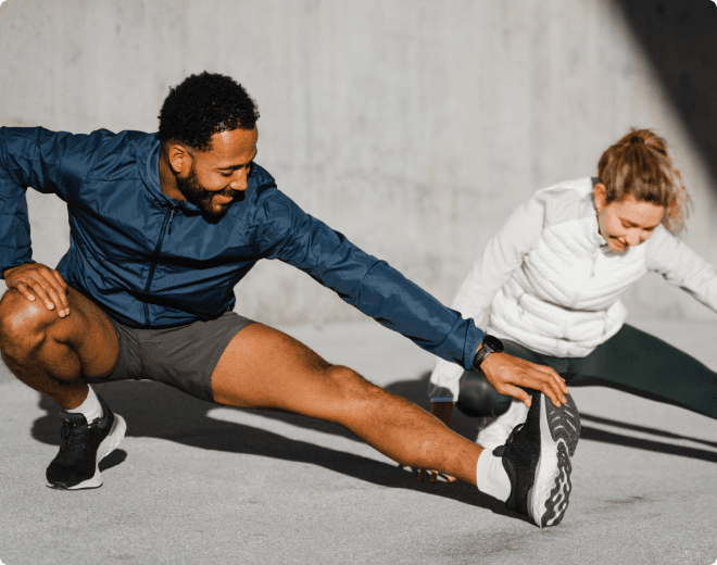 A man and a woman stretching on the floor during a workout session, both smiling and dressed in workout clothes.