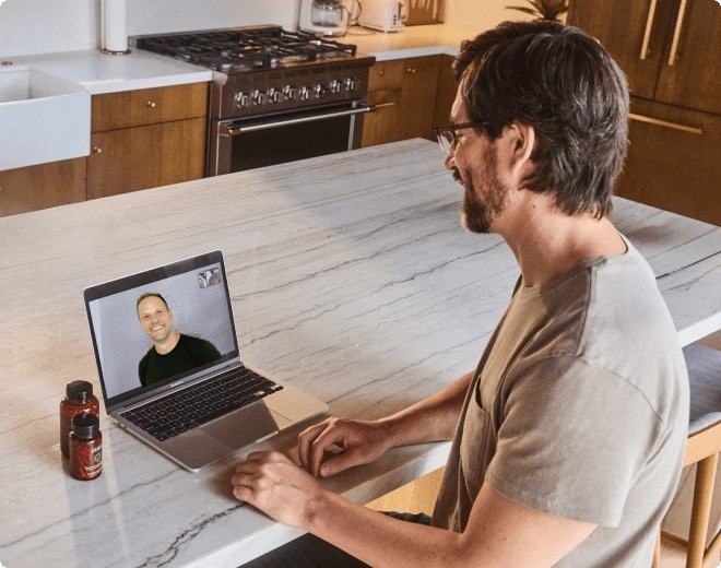 A man sitting at a kitchen counter engaged in a video call on his laptop, with another person appearing on the screen.