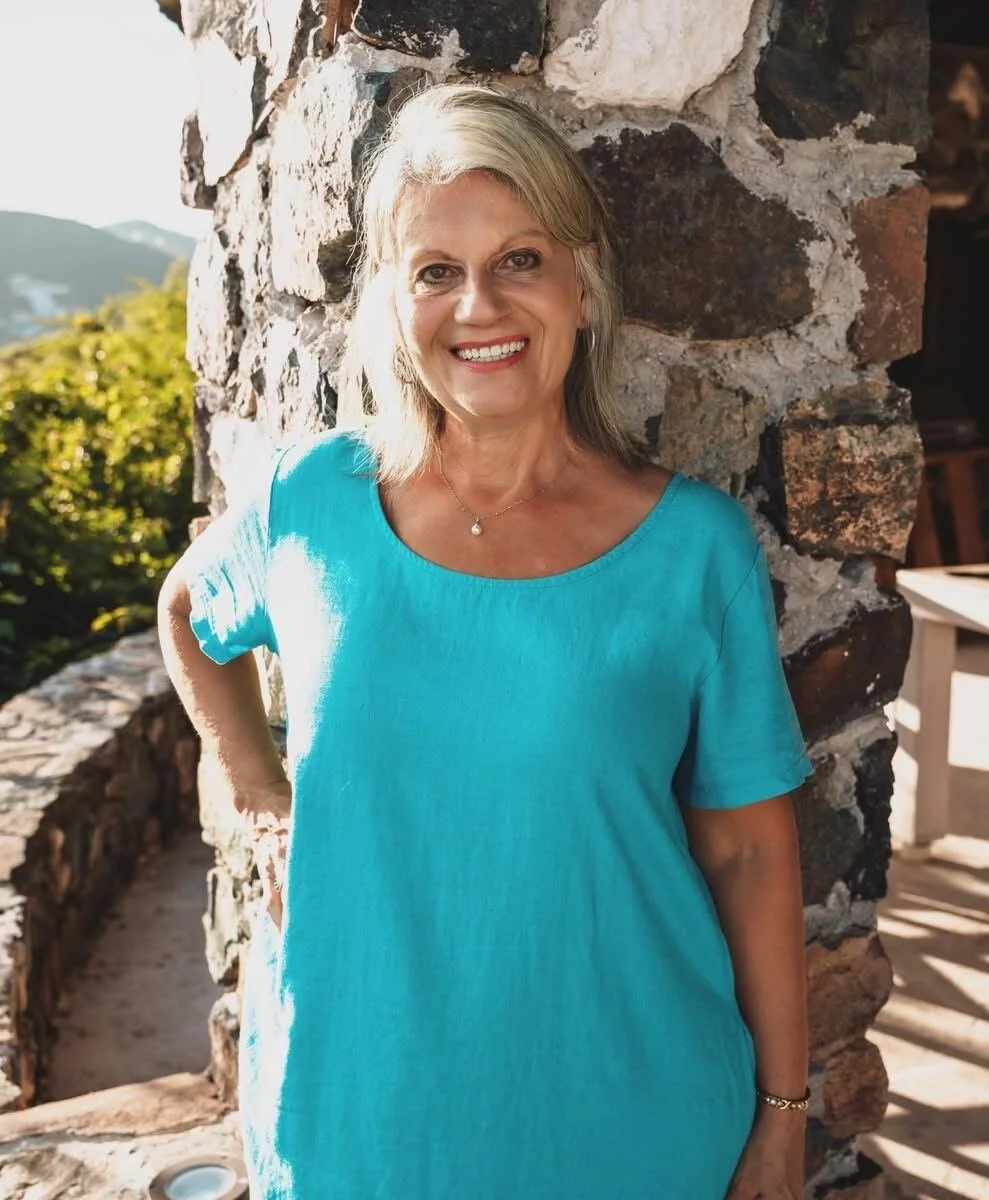 Smiling woman with gray hair wearing a turquoise dress stands next to a stone wall outdoors during daytime.