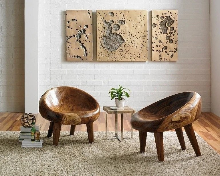 Living room with two wooden chairs, a small side table with a potted plant, a stack of books, and abstract wall art on a white brick wall.
