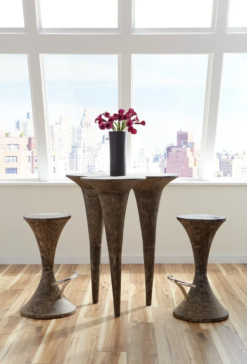 A round wooden table and two matching stools, with a black vase containing pink flowers on the table, positioned in front of large windows overlooking a cityscape.