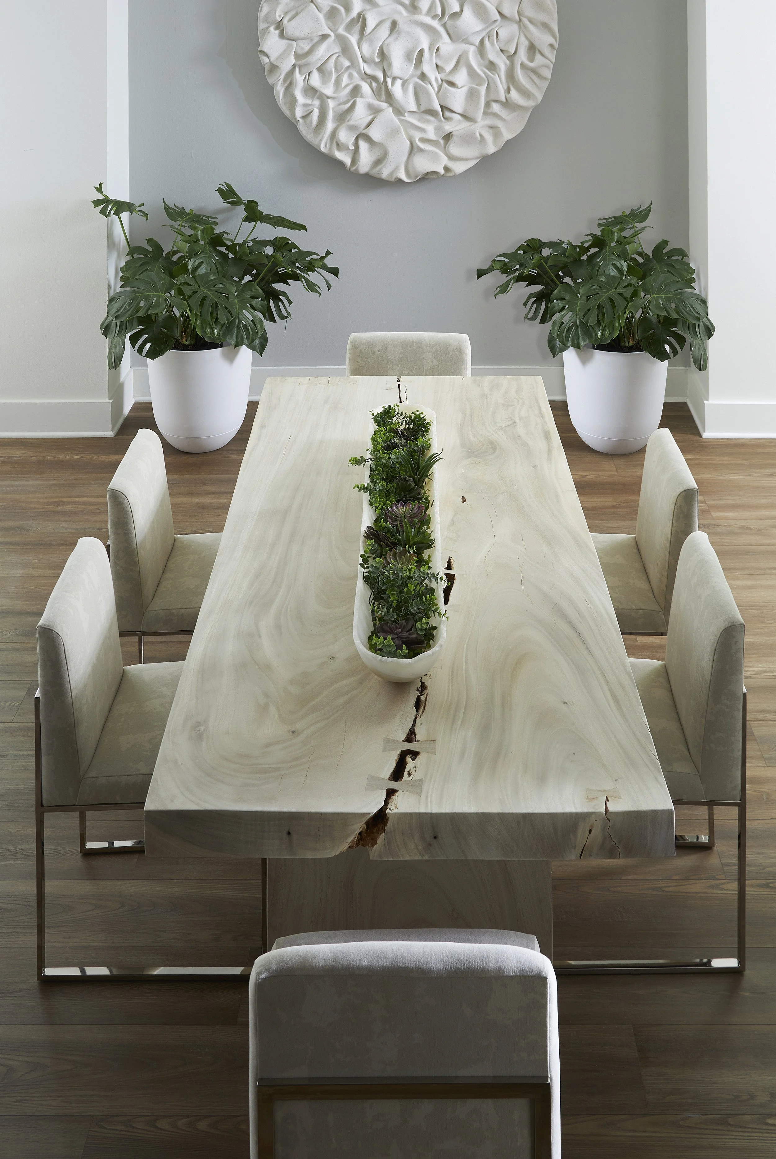 A dining room with a wooden table, surrounded by six beige upholstered chairs, featuring a long rectangular planter with succulents. Two large potted monstera plants sit on either side of the wall behind the table. A large round textured white wall art piece decorates the wall above.