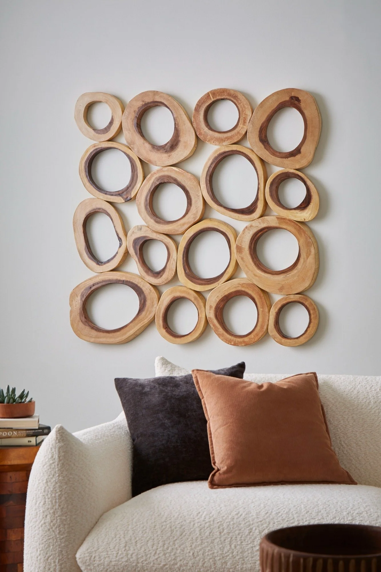 A beige upholstered armchair with a dark brown and a rust-colored throw pillow, a small side table with a potted plant, books, and a brown ceramic vase, against a light gray wall adorned with a wooden abstract wall art piece.