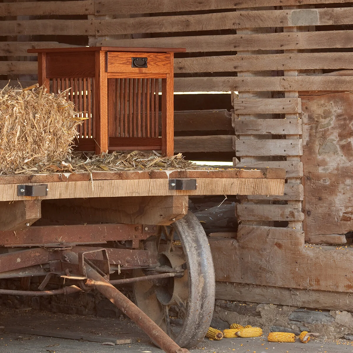 A small wooden table or cabinet with a single drawer, placed on a rustic wooden platform, with dried corn husks nearby, set against a weathered wooden wall with gaps between the planks.