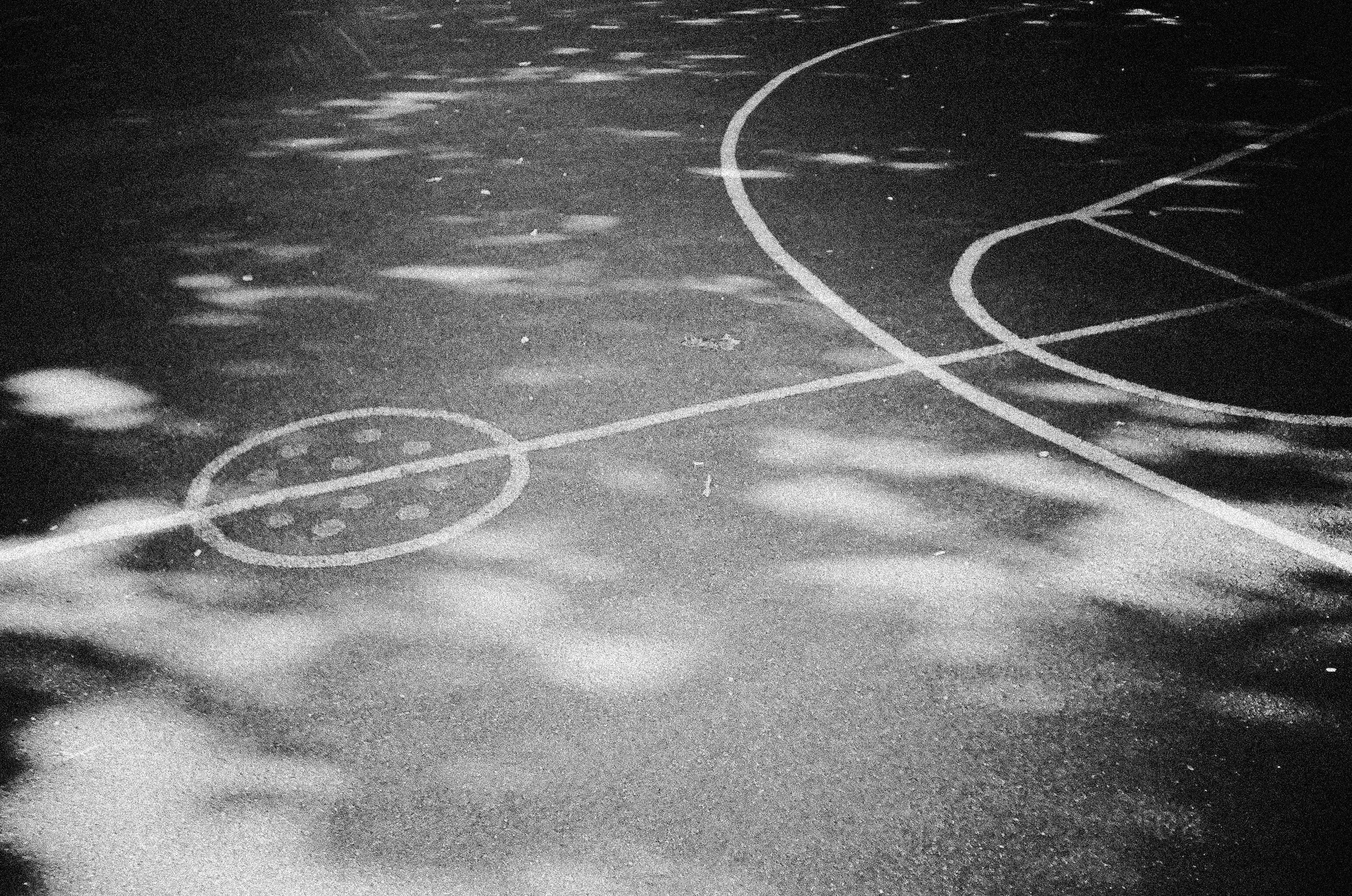 A black and white nighttime image of a basketball court with visible court markings, illuminated by an overhead light, creating shadows and reflections on the surface.