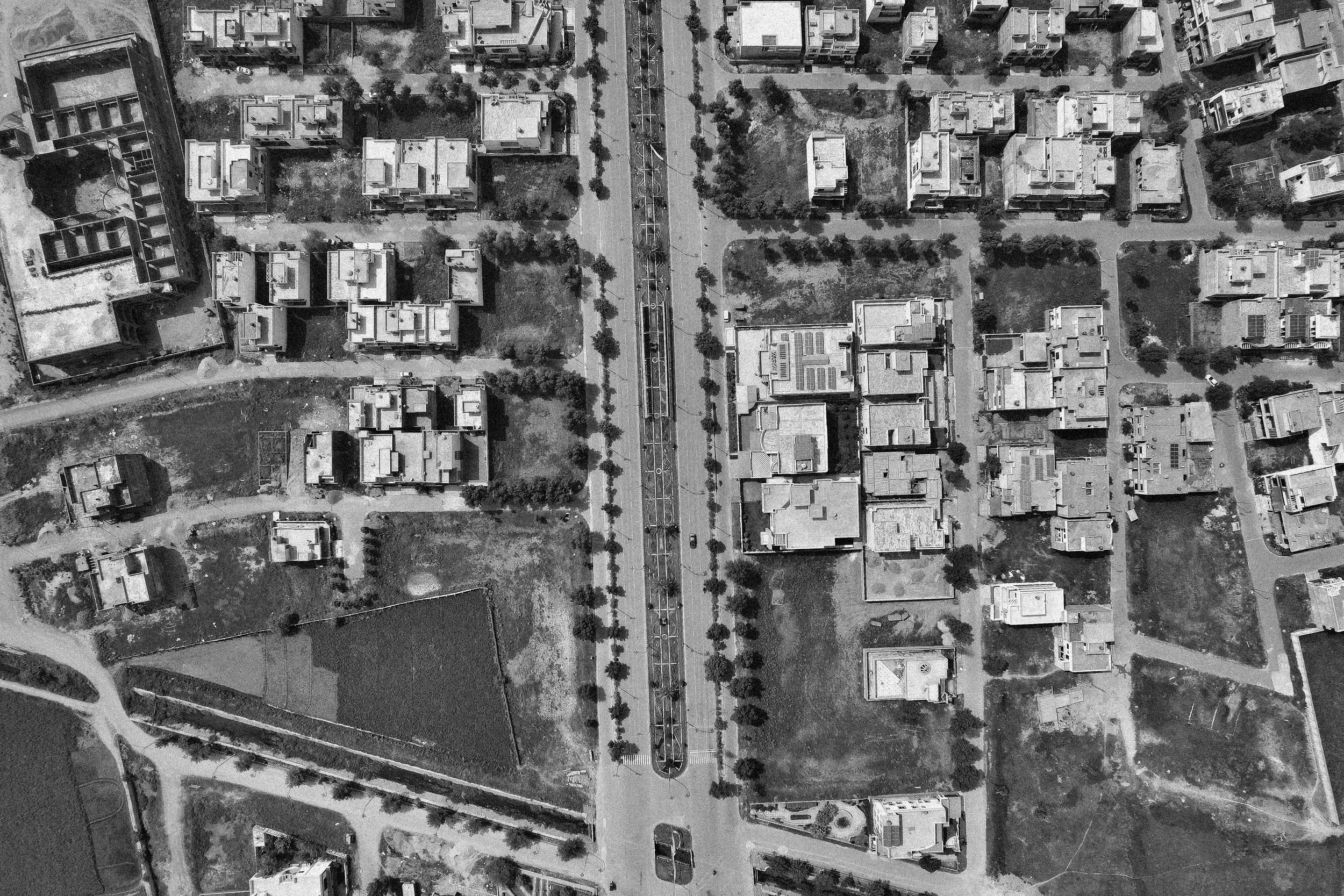 Aerial view of a cityscape showing multiple buildings, roads, trees, and open spaces in black and white.