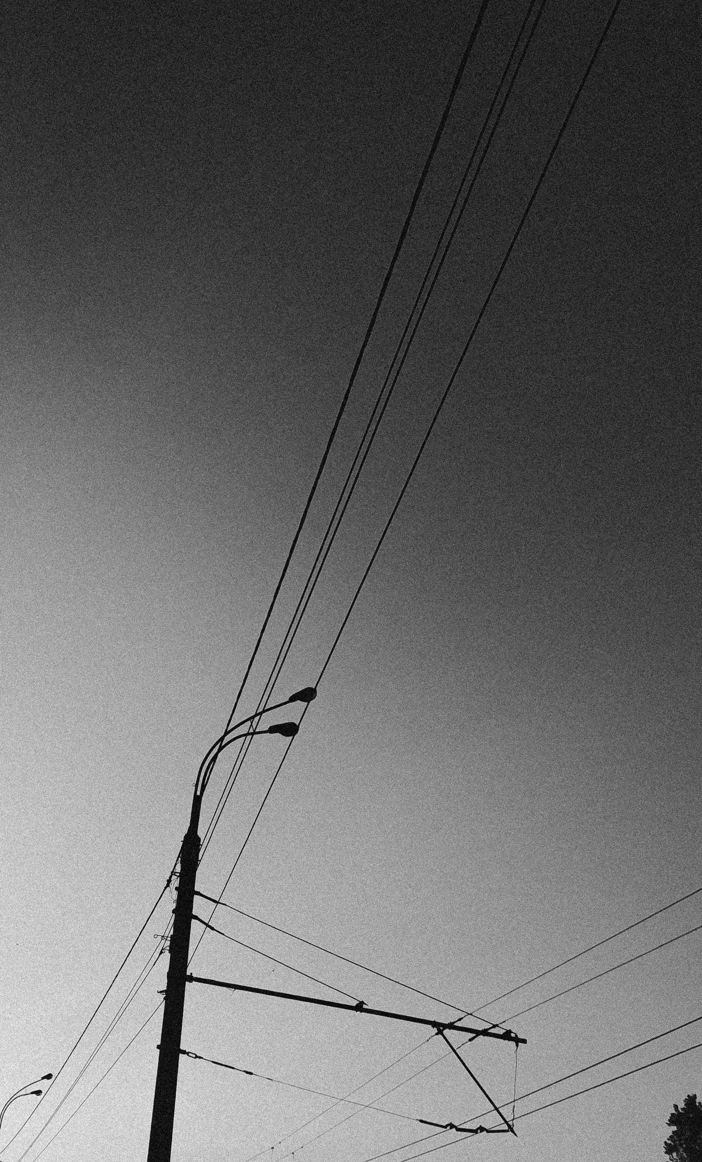 Black and white photo of a utility pole with multiple power lines and streetlights against a sky that is darker at the top and lighter at the bottom.