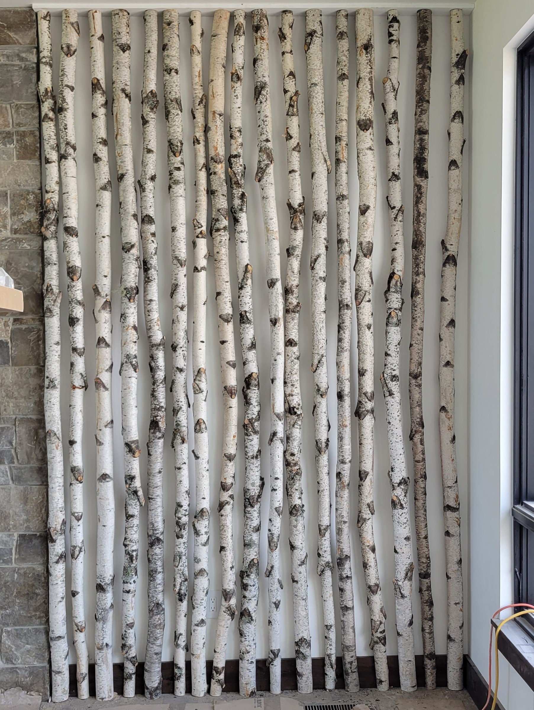 A wall with vertical birch log pieces arranged in a decorative pattern indoors, next to a stone wall and near a window with electrical wires and outlet nearby.