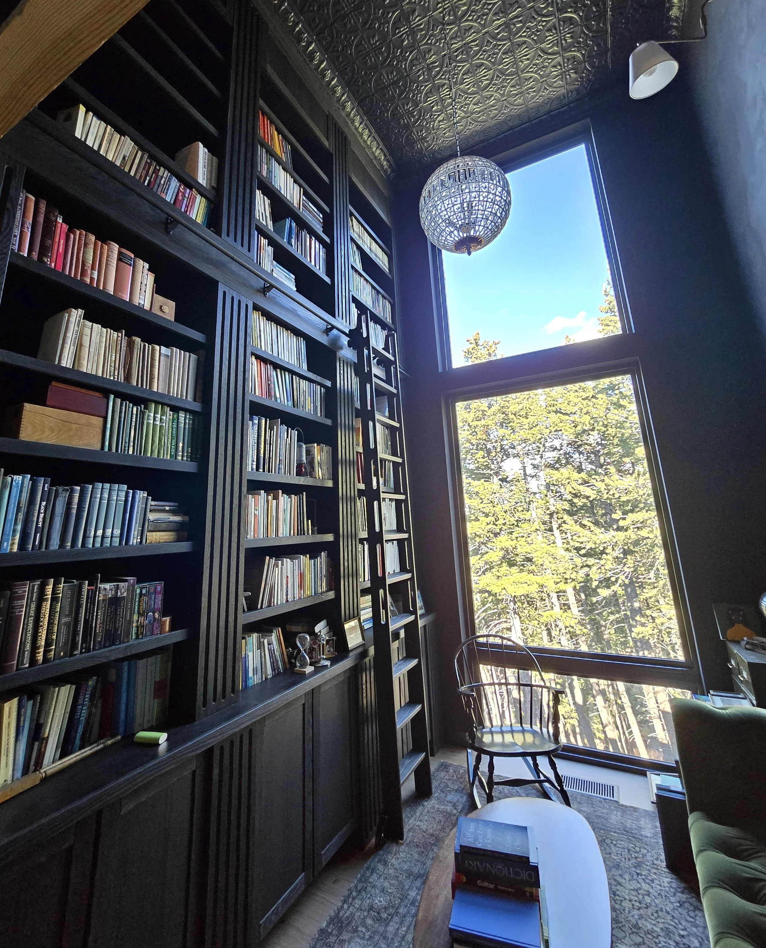 A cozy library or study room with tall, dark bookshelves filled with books, a large window showing trees and blue sky outdoors, a white table holding stacked books, a green armchair, a patterned rug, and a decorative ceiling with an intricate design. A spherical hanging light fixture is also visible.