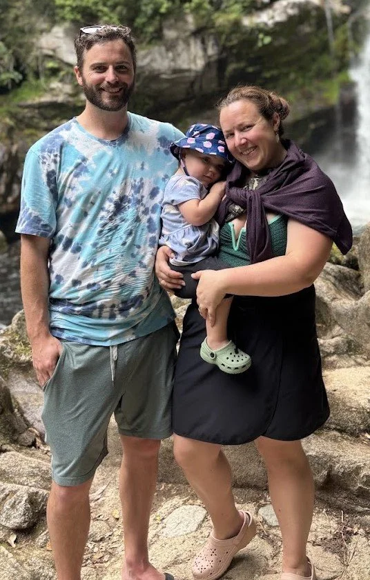 A smiling family of three outdoors near a waterfall, with the man on the left, the woman on the right holding their young child in her arms, all standing on rocks surrounded by greenery.