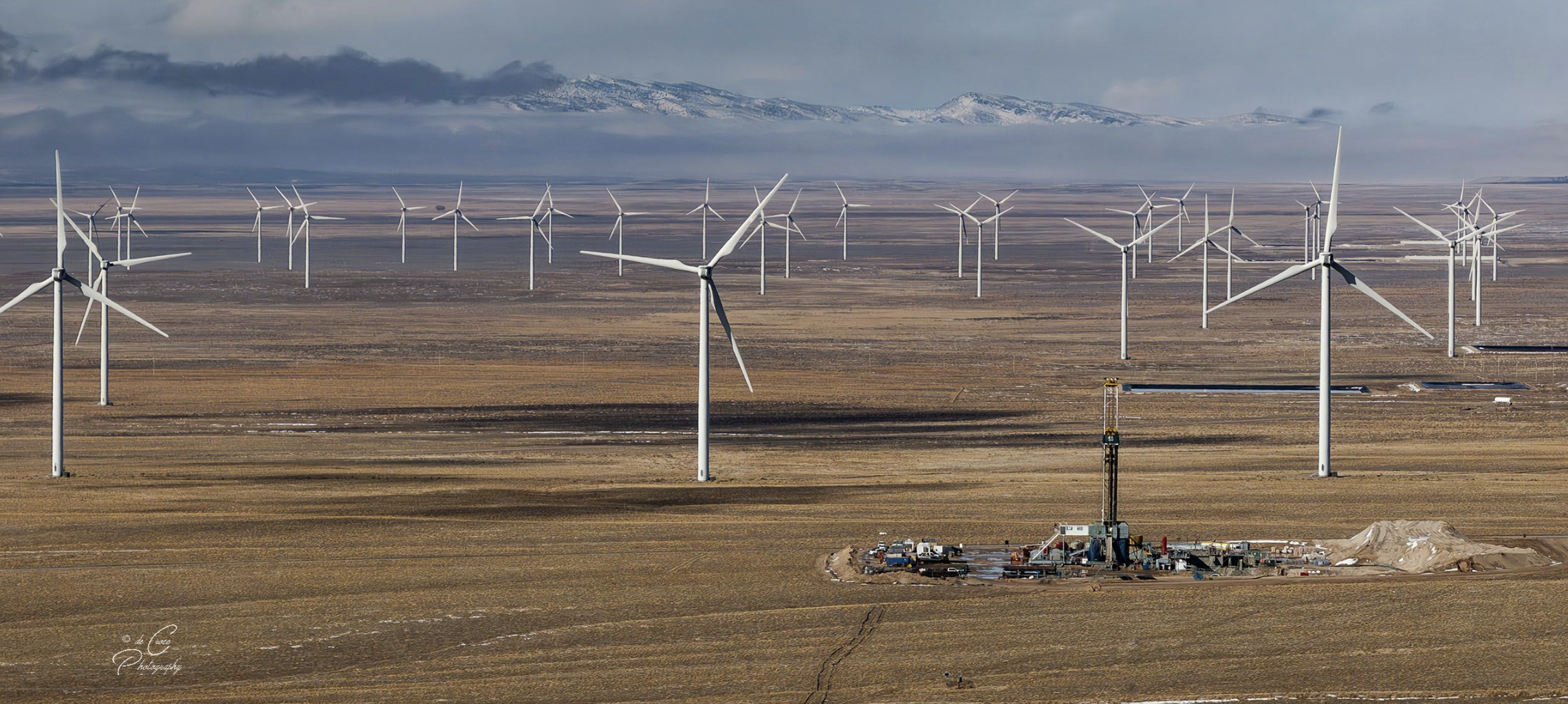 A large field with numerous wind turbines under a cloudy sky, with mountains in the background, and a construction site with equipment and vehicles in the foreground.