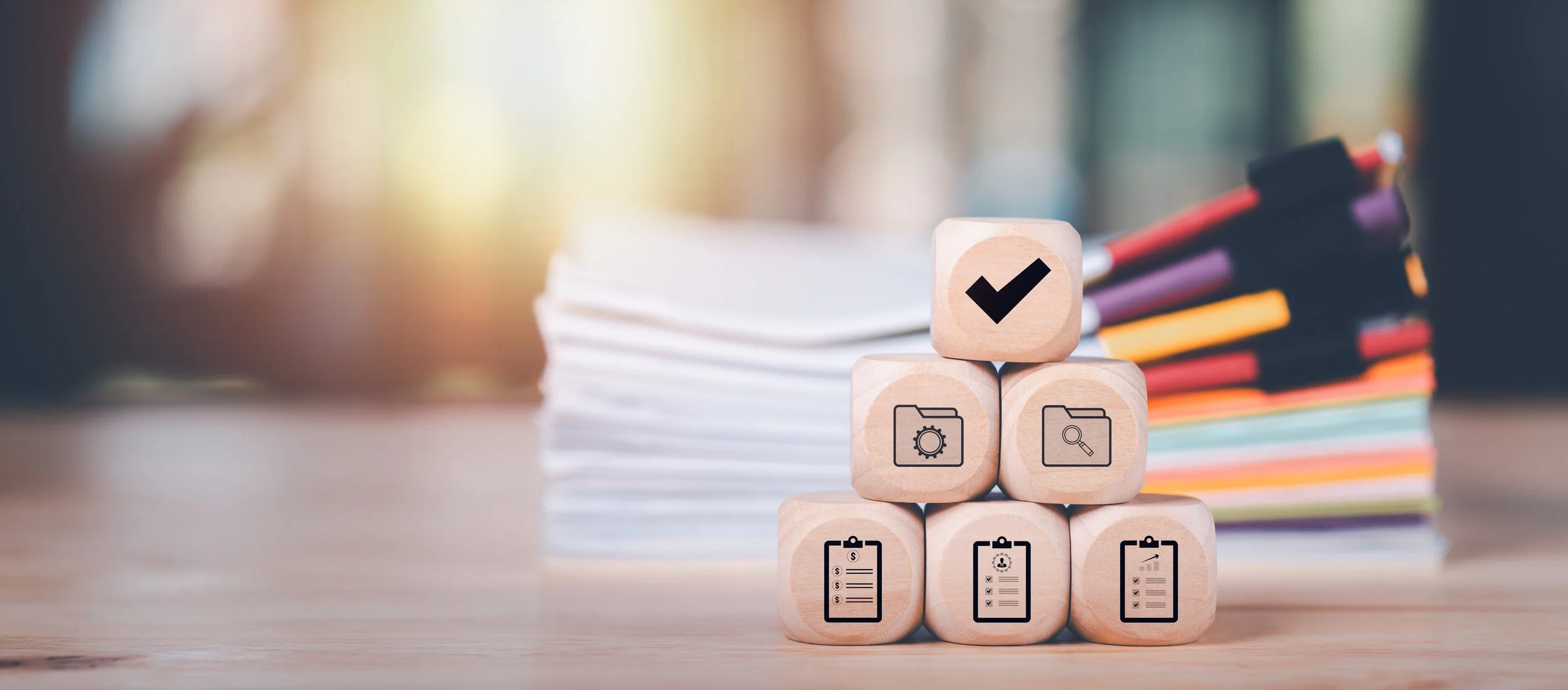 Wild Goose Studios - exceptional execution - Stacked wooden blocks with icons of a checklist, camera settings, folder with gear, and magnifying glass, topped with a checkmark symbol, with books and papers in the background.