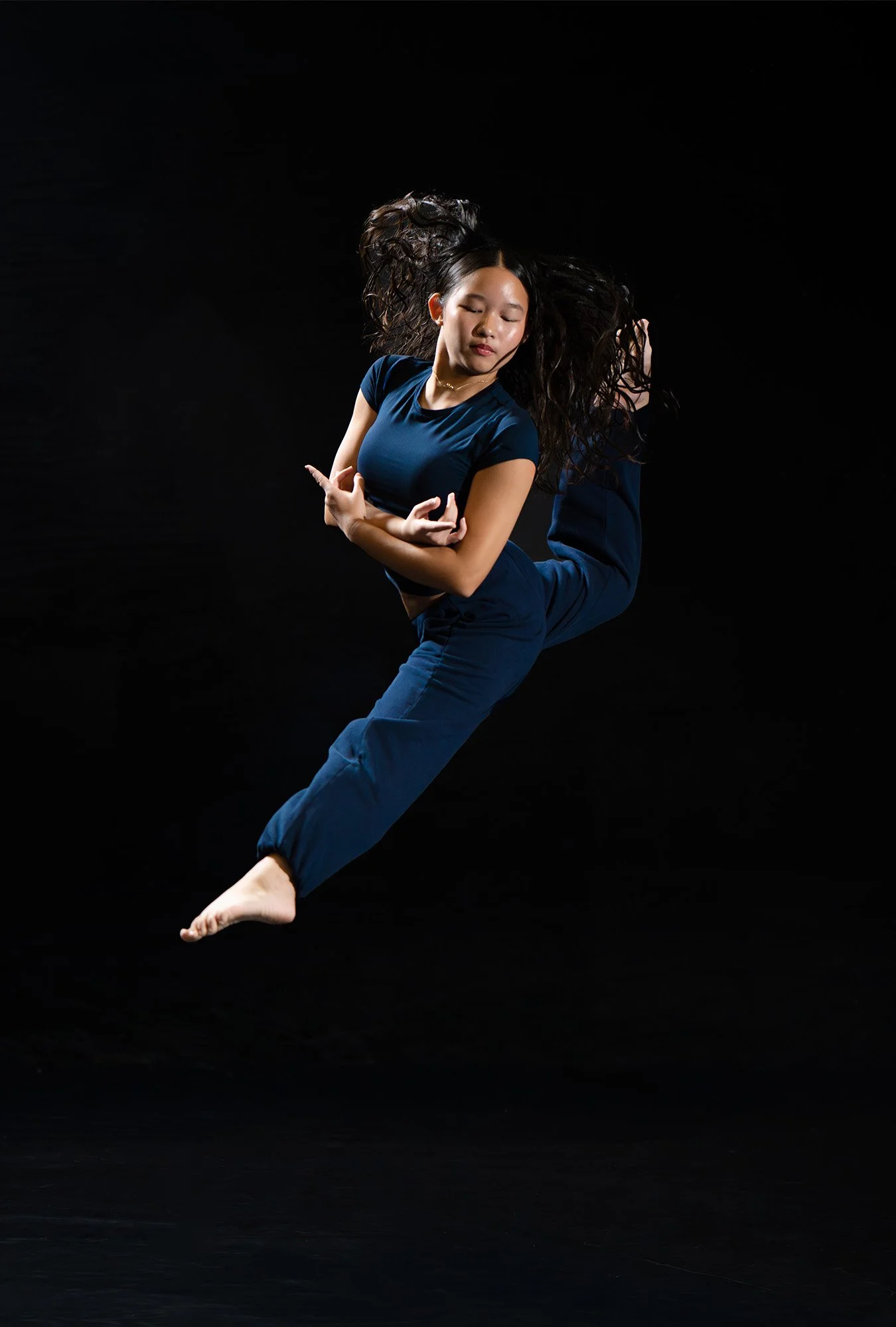 A young woman with long dark hair in a blue outfit is performing a dance move against a black background, with her eyes closed and arms crossed in front of her chest.
