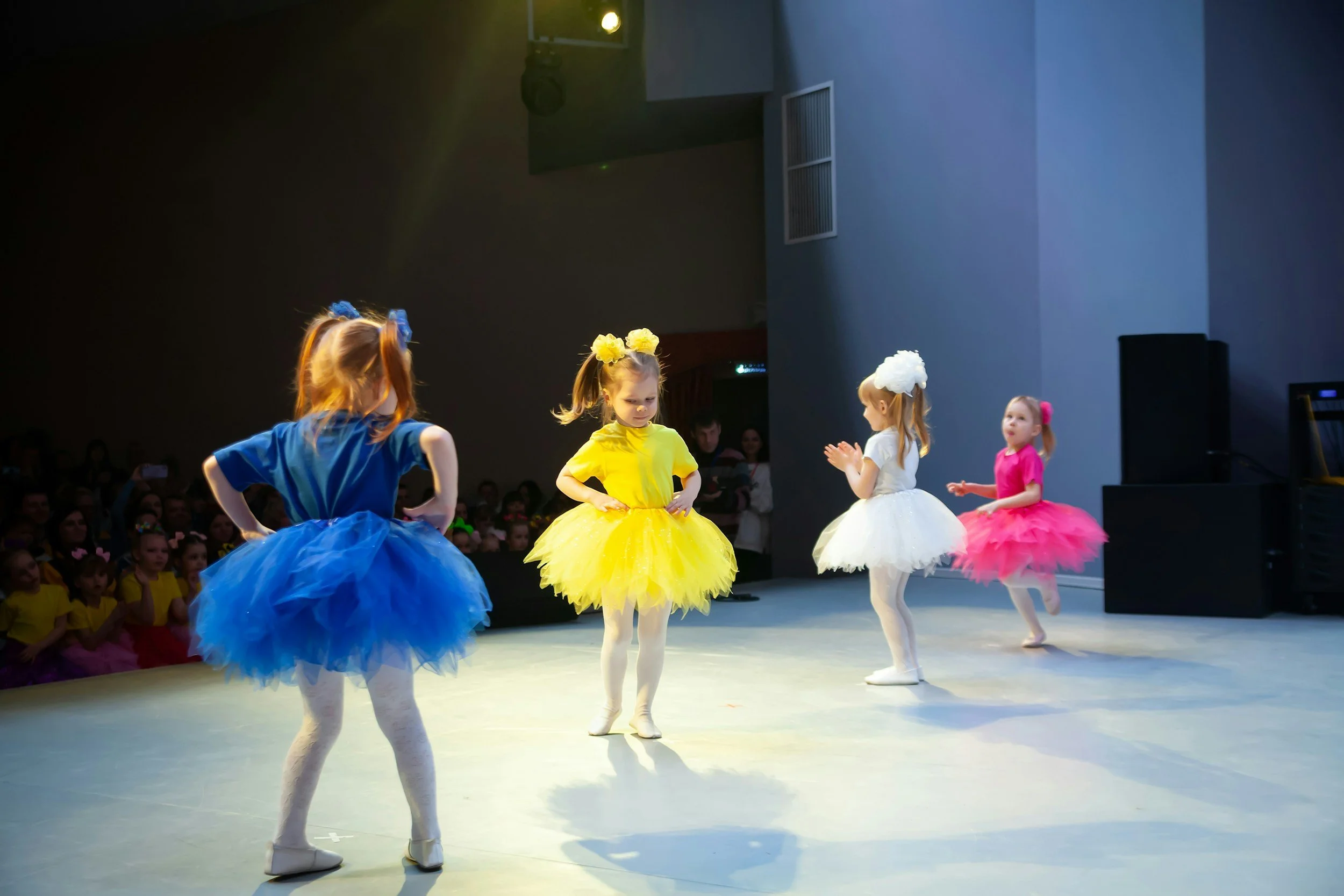Children in colorful tutu dresses performing ballet on stage during a dance performance, with audience members watching in the background.
