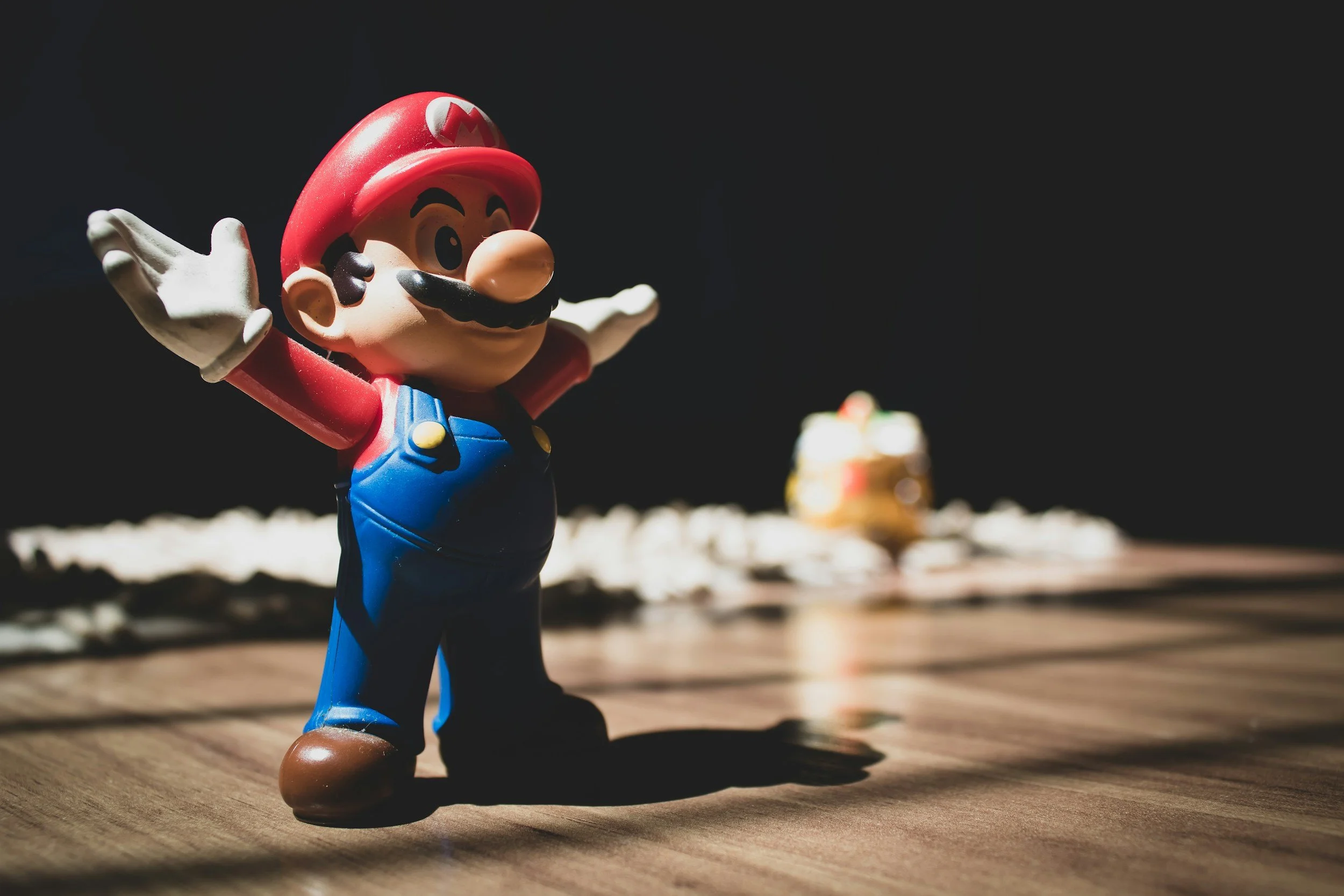 Close-up of a Mario figurine with arms spread, standing on a wooden surface, with a blurred background.