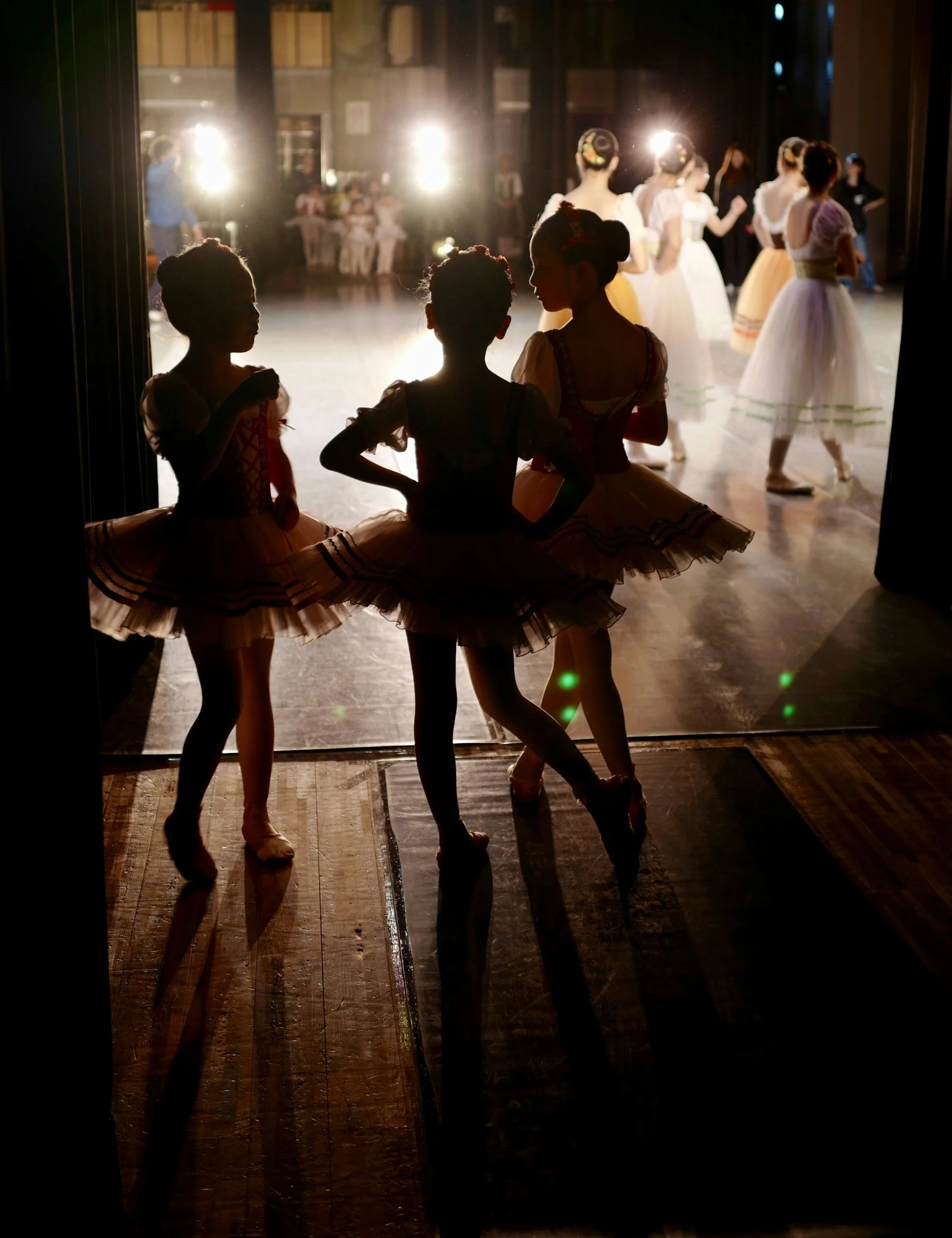 Backstage view of ballet dancers in tutus waiting to go on stage in a dimly lit theater with bright stage lights shining through the curtain, illuminating some dancers' silhouettes.